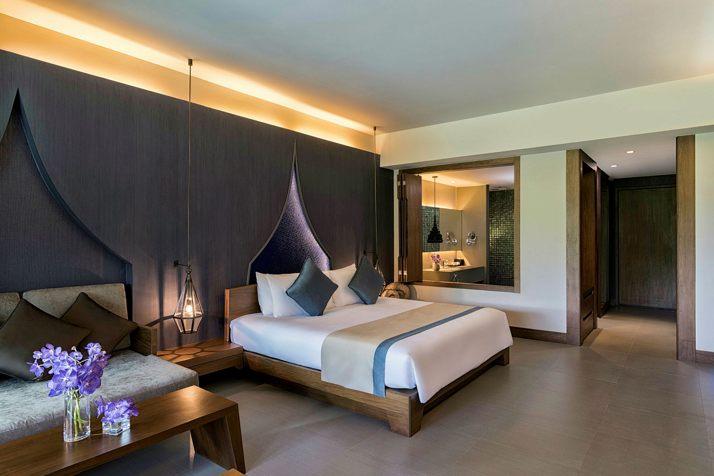 Et hotellrom med moderne innredning. Sengen er dekket med hvite laken og blå dekorative puter. Ved siden av sengen står et bord med blomster i en glassvase. Et mørkt teppe vises bak sengens hodegavl. (AI-generert tekst)