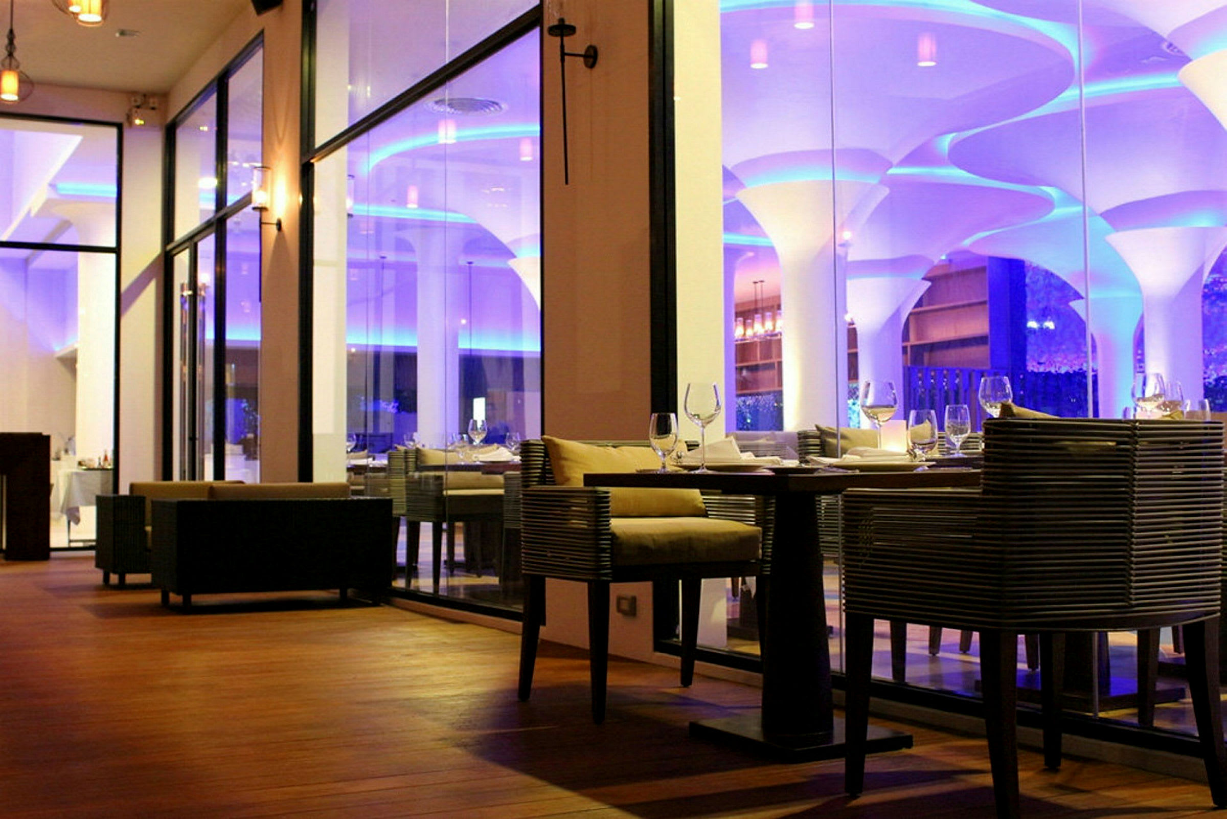 Interiør av en elegant restaurant med glassvegger og moderne møbler, belysning i blått og varmt lys. (AI-generert tekst)
