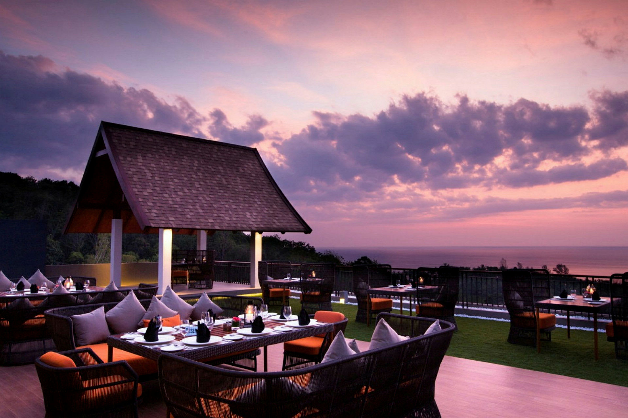 En utendørsrestaurant med bord og stoler under en lilla og rosa himmel i solnedgangen, med utsikt over havet. (AI-generert tekst)