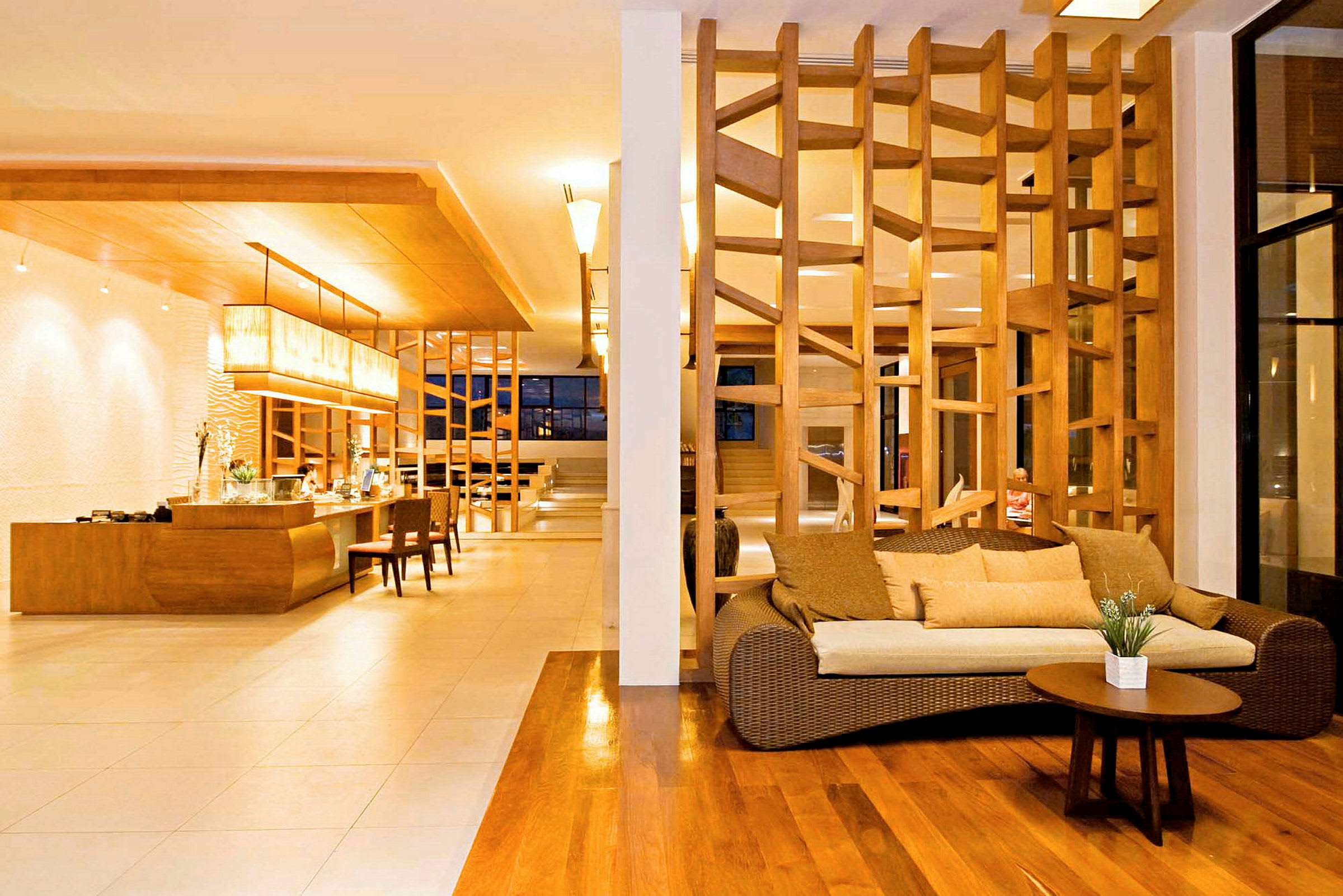 En hotellobby med tregulv, en sofa med puter og et lavt bord i forgrunnen, samt en designet tretillatelse som avdeling. Bakgrunnen viser resepsjonsområdet med belysning ovenfor. (AI-generert tekst)