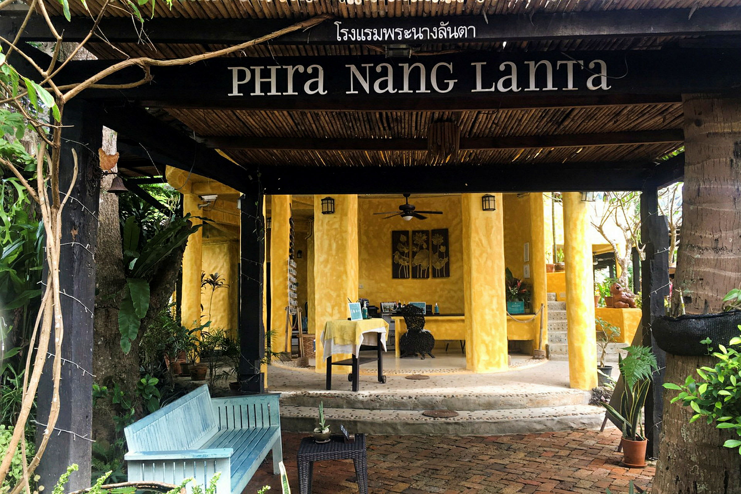 Inngangen til et hotell med navnet Phra Nang Lanta, med gule vegger og tredetaljer, omgitt av grønt. (AI-generert tekst)