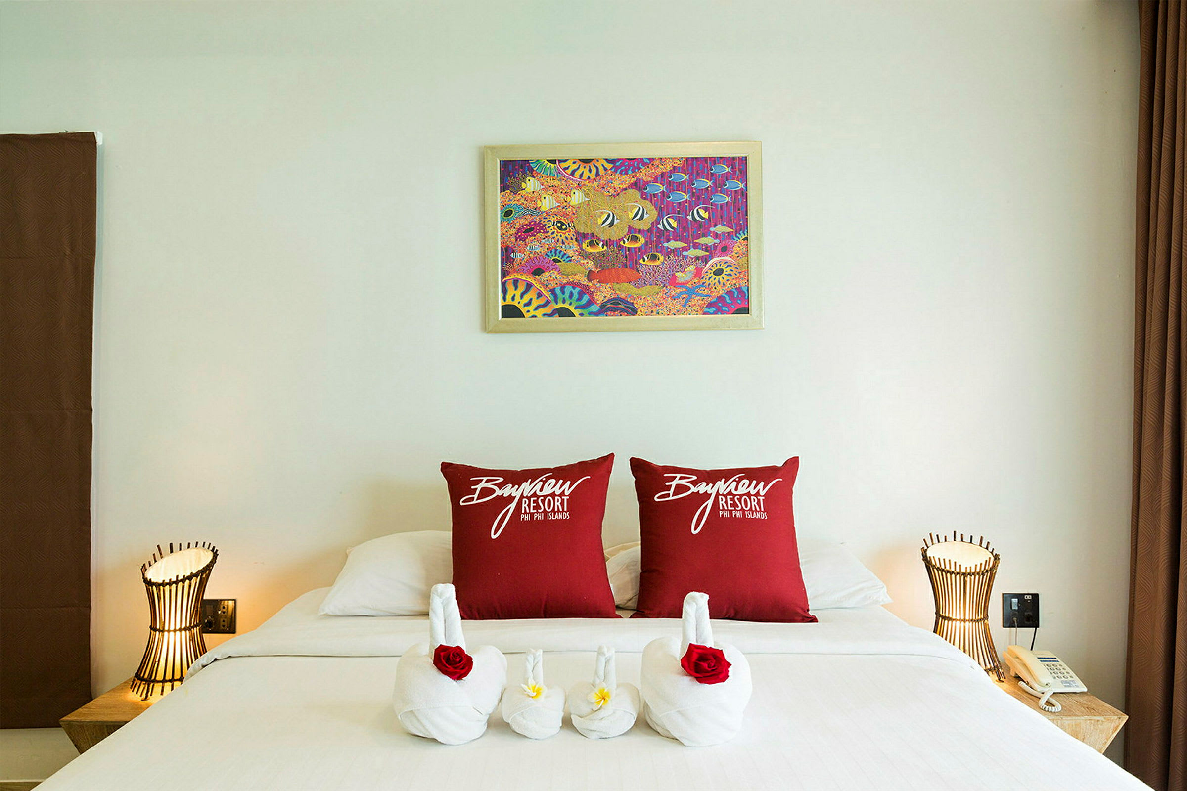 Et hotellrom med en dekorert seng. To røde puter med teksten «Bayview Resort» er plassert på sengen, og håndduker er arrangert som svaner med blomster. Det er en lampe og en telefon på et nattbord. (AI-generert tekst)