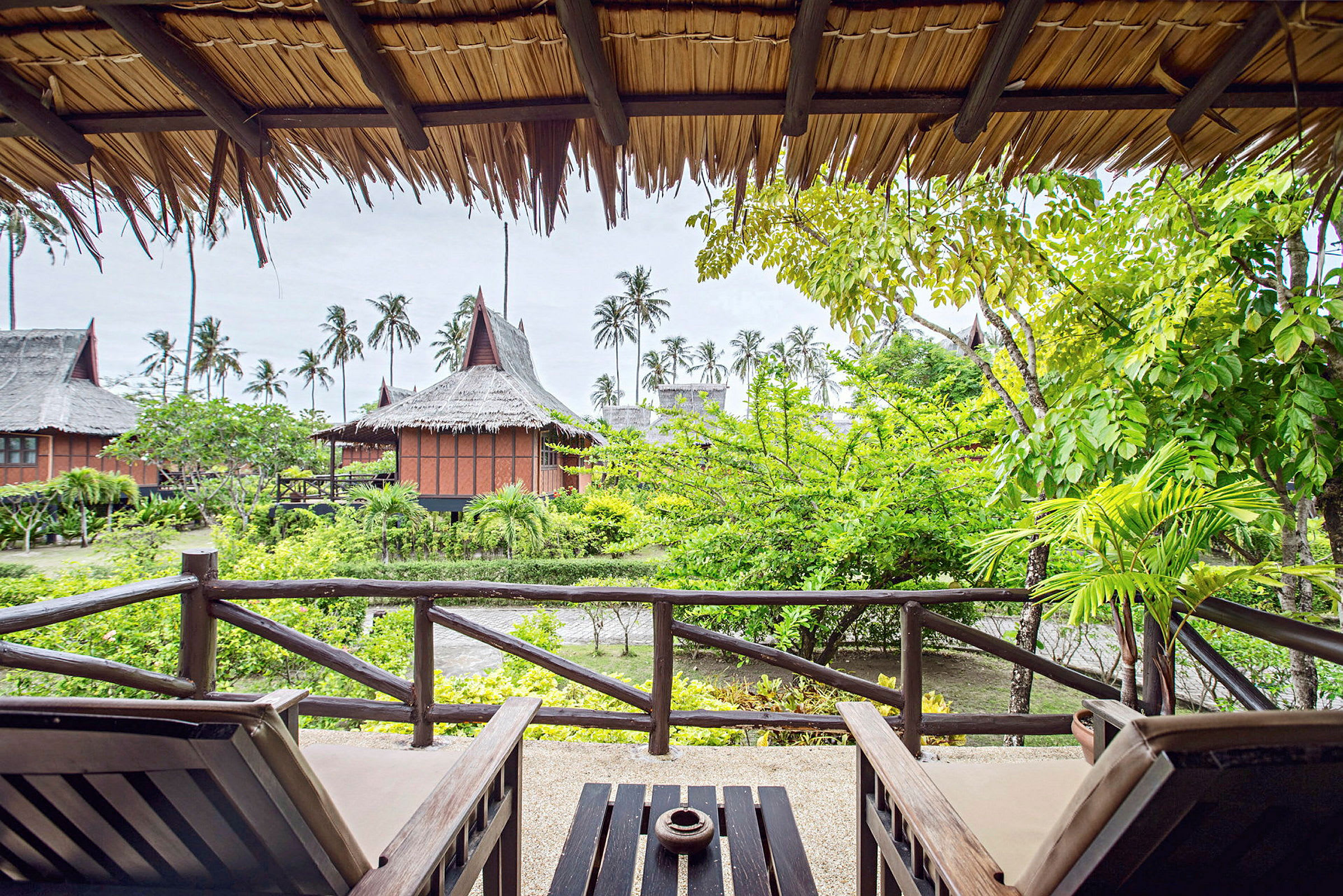 Utsikt fra en veranda med to solsenger mot en hage med grønne planter og tradisjonelle bygninger med stråtak. (AI-generert tekst)