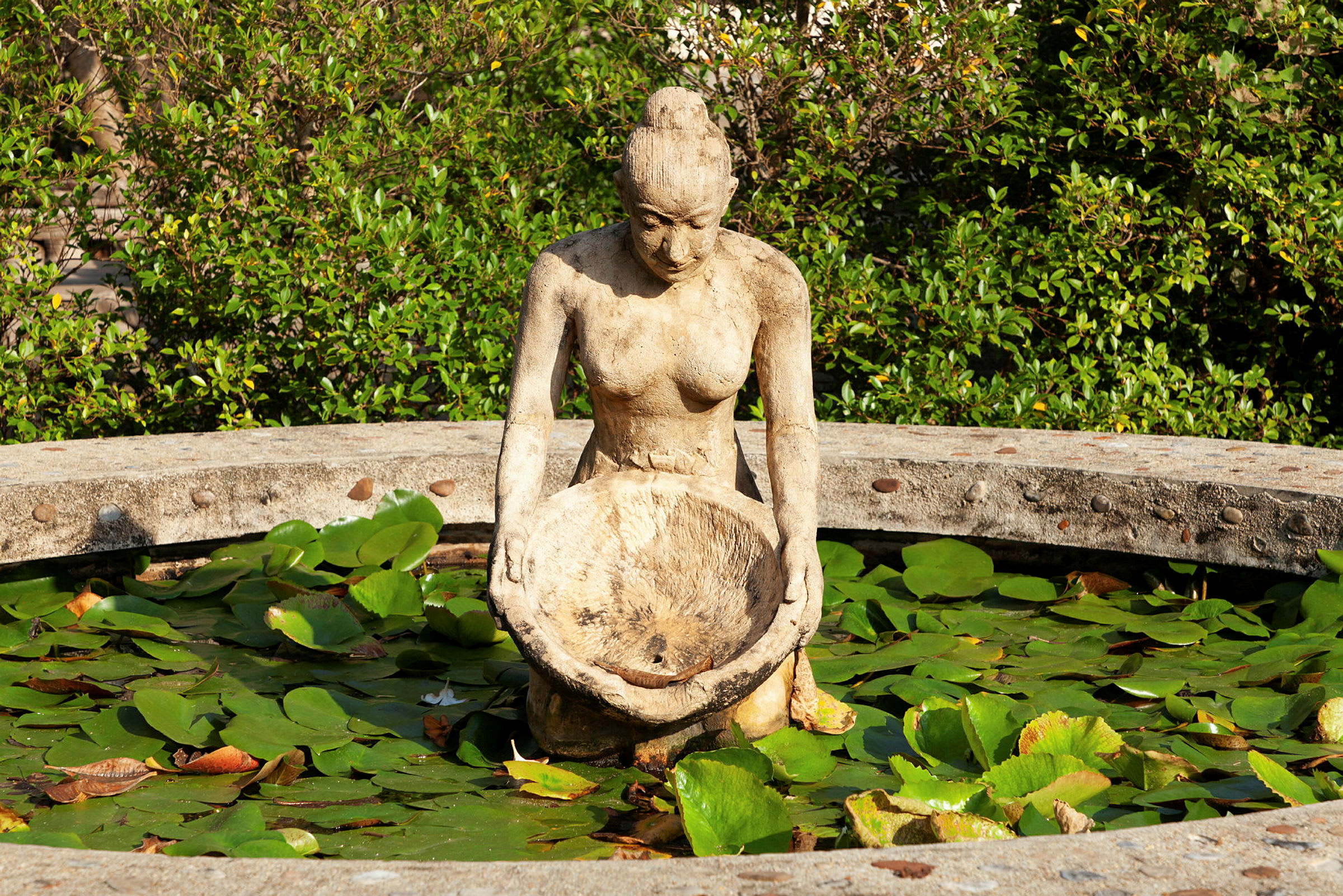 Statue av en kvinne som holder en bolle, omgitt av et sirkulært vannfyllt kar med vannliljeblader. (AI-generert tekst)