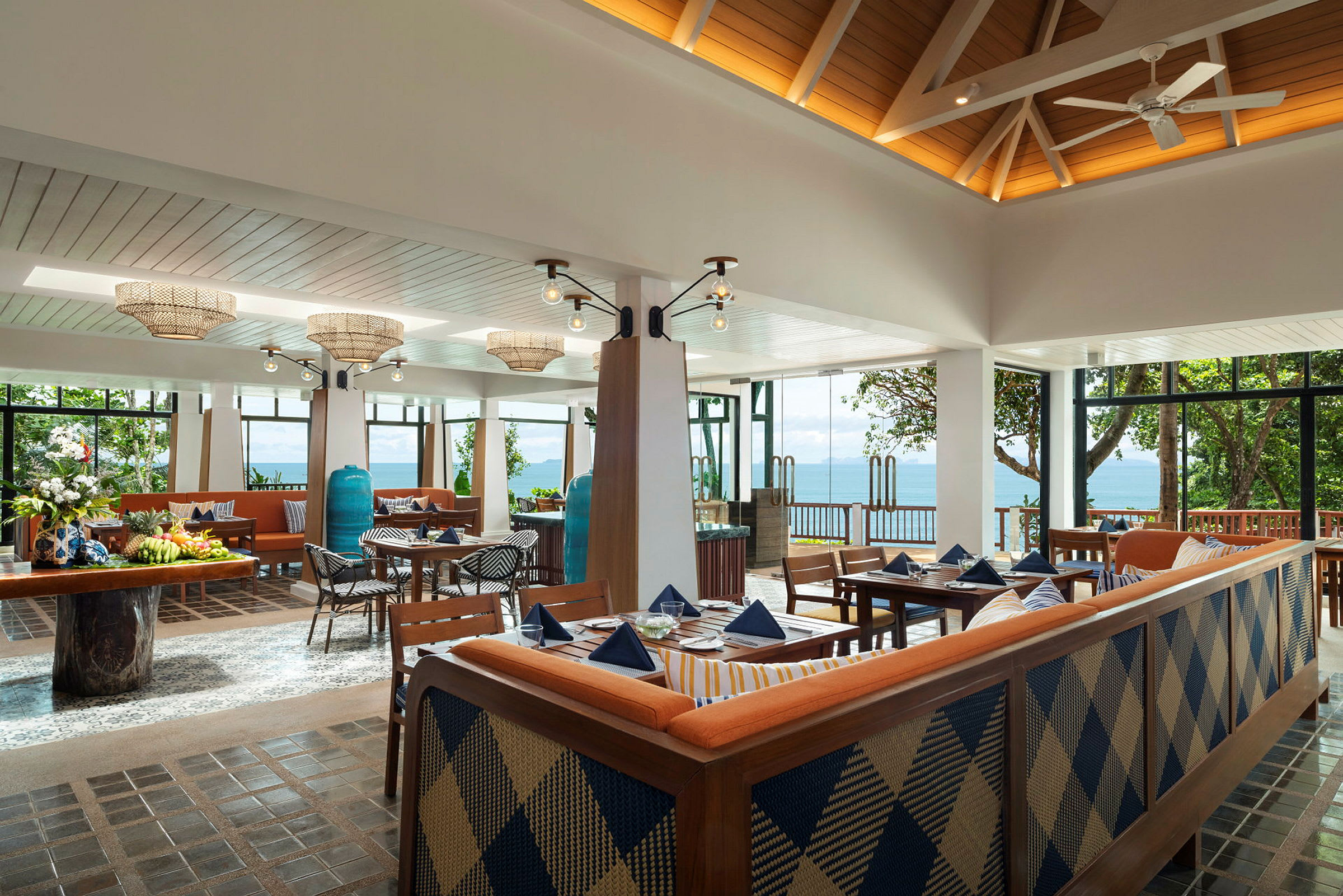 Et luftig og lyst restaurantområde med store vinduer mot havet. Møblene består av bord med blå duker, trestoler og sofaer. (AI-generert tekst)