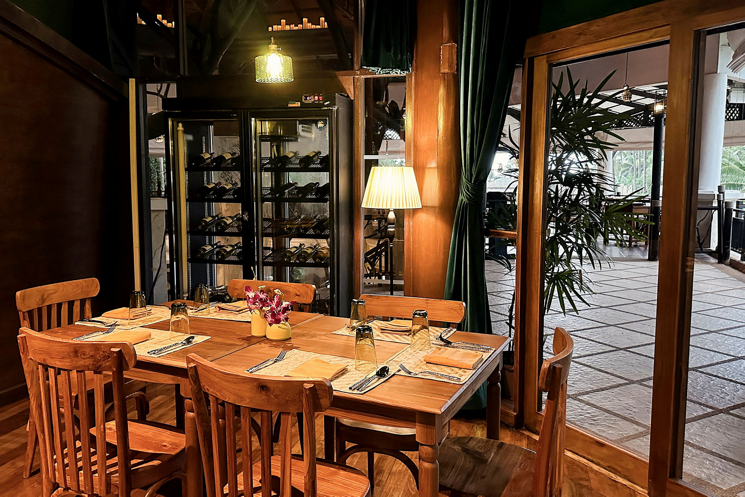 Et trebord med trestoler er dekket for fire personer i en restaurantmiljø. Vinreoler og grønne gardiner er synlige i bakgrunnen. (AI-generert tekst)