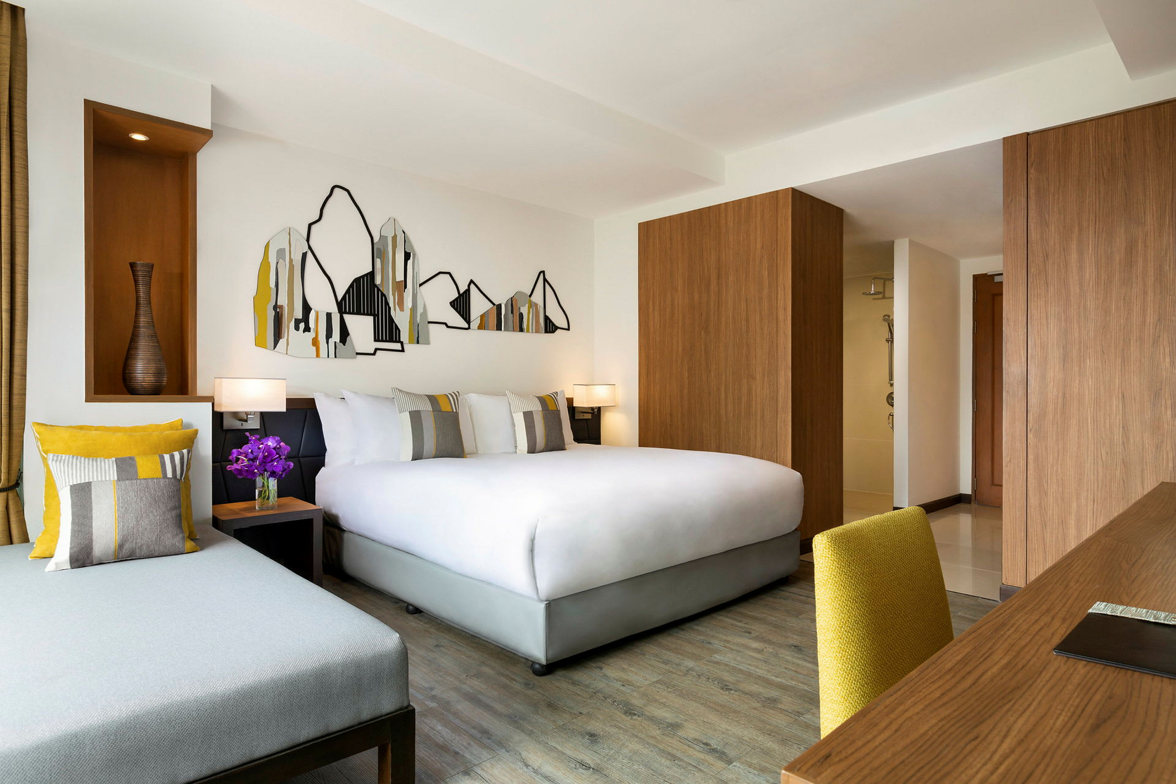 Et moderne hotellrom med en dobbeltseng, en sofa og et skrivebord med gul stol. Veggdekorasjoner med fjellmotiv i abstrakt stil. (AI-generert tekst)