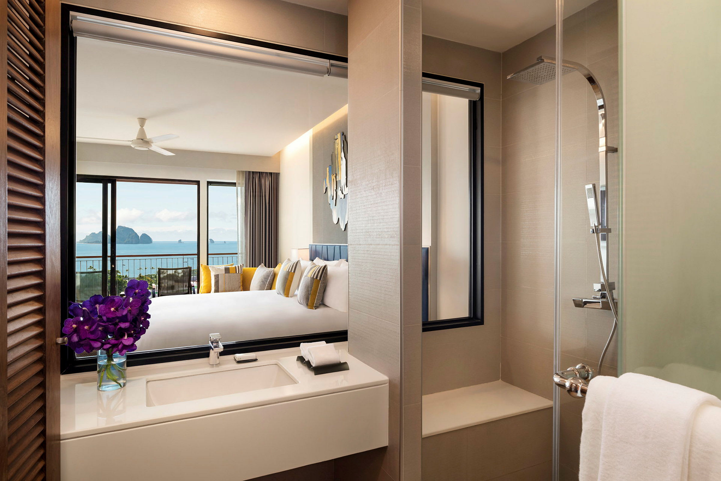 Et moderne hotellrom med utsikt over havet. Bildet viser badet med en vask og blomster, samt en dusj i forgrunnen og et tilknyttet soverom i bakgrunnen med stor seng og havutsikt. (AI-generert tekst)
