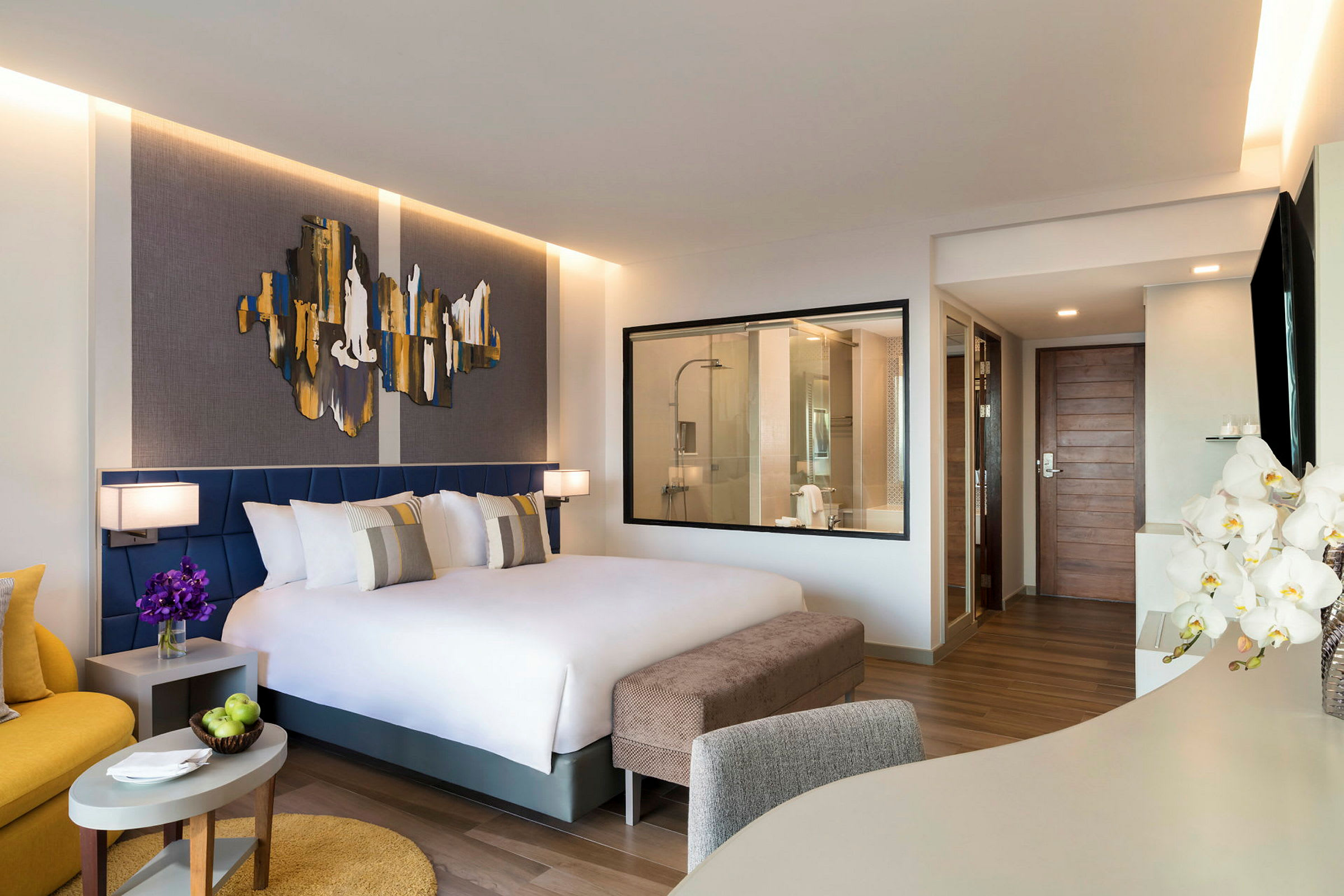Et moderne hotellrom med en stor seng, to nattbord med lamper, et vindu inn til badet og en sittegruppe i forgrunnen. (AI-generert tekst)