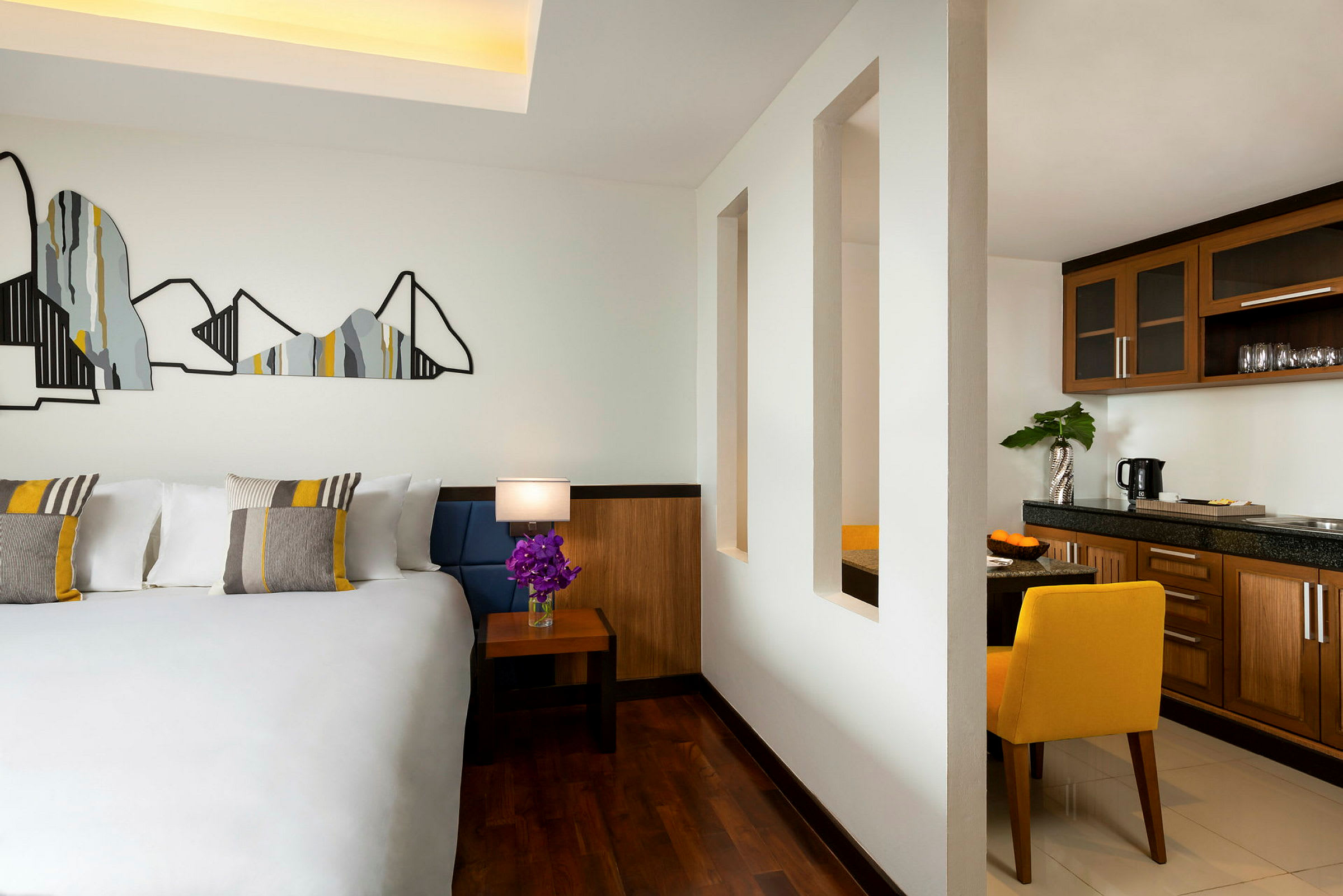 Et hotellrom med en stor seng med dekorative puter, et sidebord med en lampe og en blomsterbukett, og en åpen del mot en kjøkkenkrok med treinnredning. (AI-generert tekst)