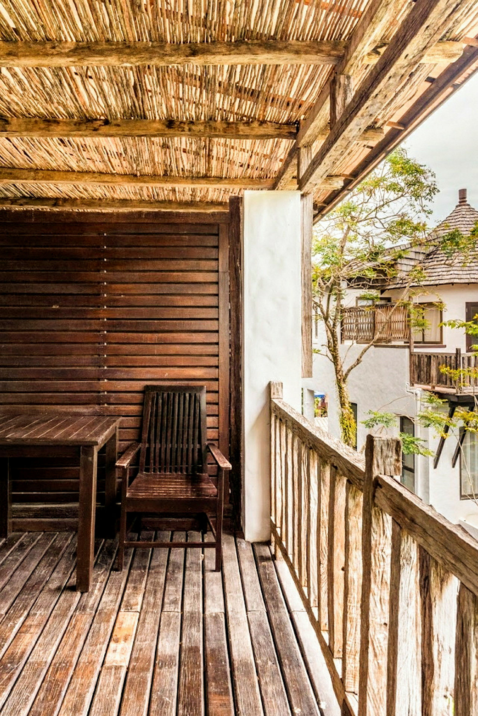 Bildet viser en veranda med tretak og tregulv. På verandaen er det en trestol og et trebord. Utsikt mot andre hus med tredetaljer og grønne omgivelser. (AI-generert tekst)