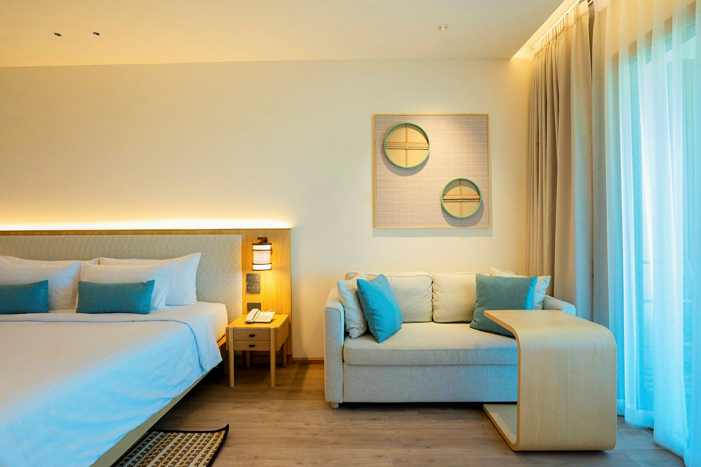 Et hotellrom med en dobbeltseng, en sofa med puter og et lite trebord. Lyse gardiner dekker et vindu i bakgrunnen. (AI-generert tekst)