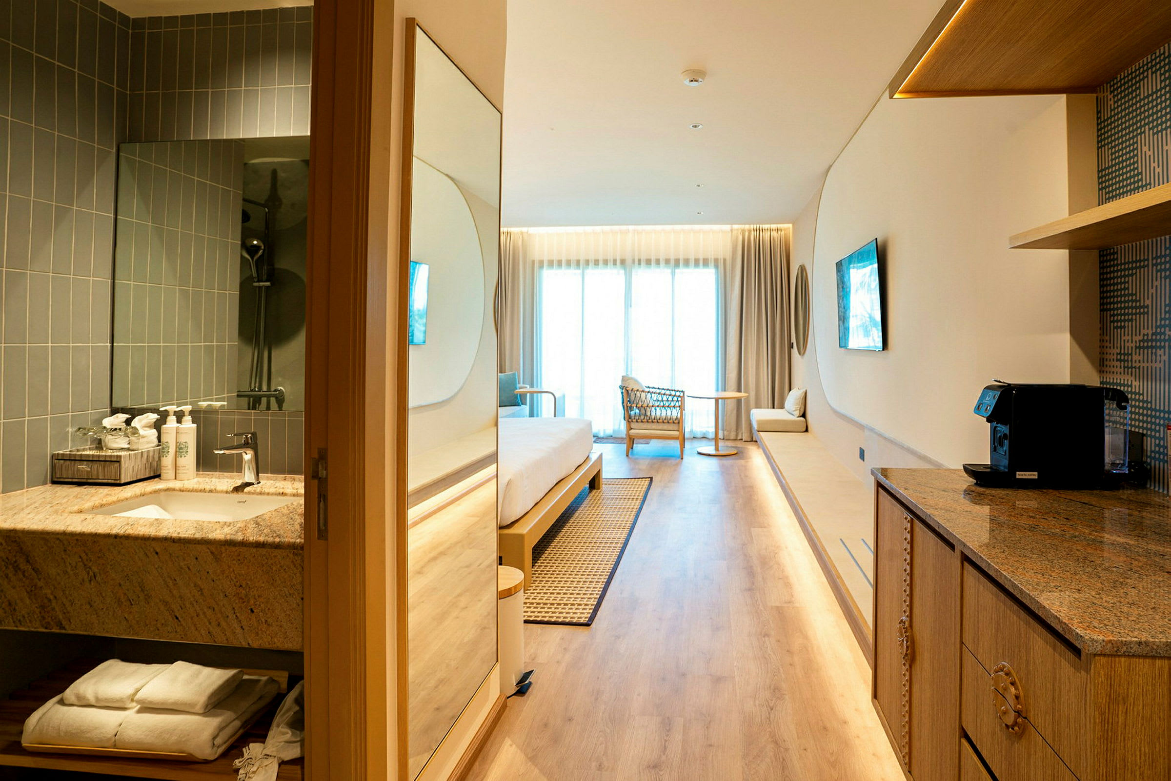 Et moderne hotellrom med lyst interiør, inkludert seng, lenestol, lite bord og balkong. Et bad og en kjøkkendel er synlig i forgrunnen. (AI-generert tekst)