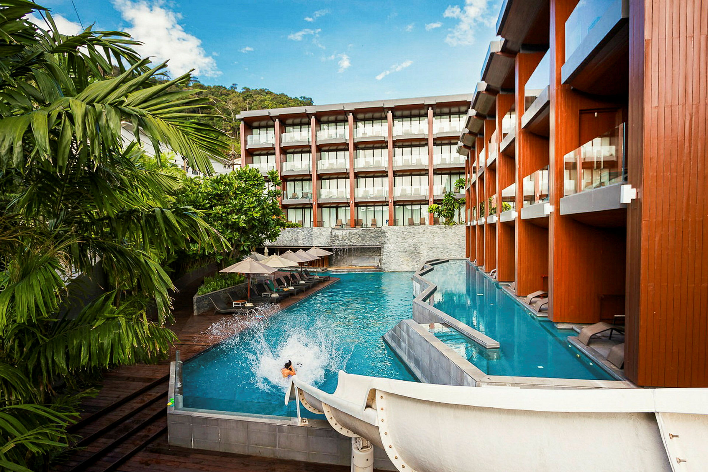 Et basseng ved et moderne hotell med solsenger og parasoller samt en vannsklie som leder ned i vannet. (AI-generert tekst)