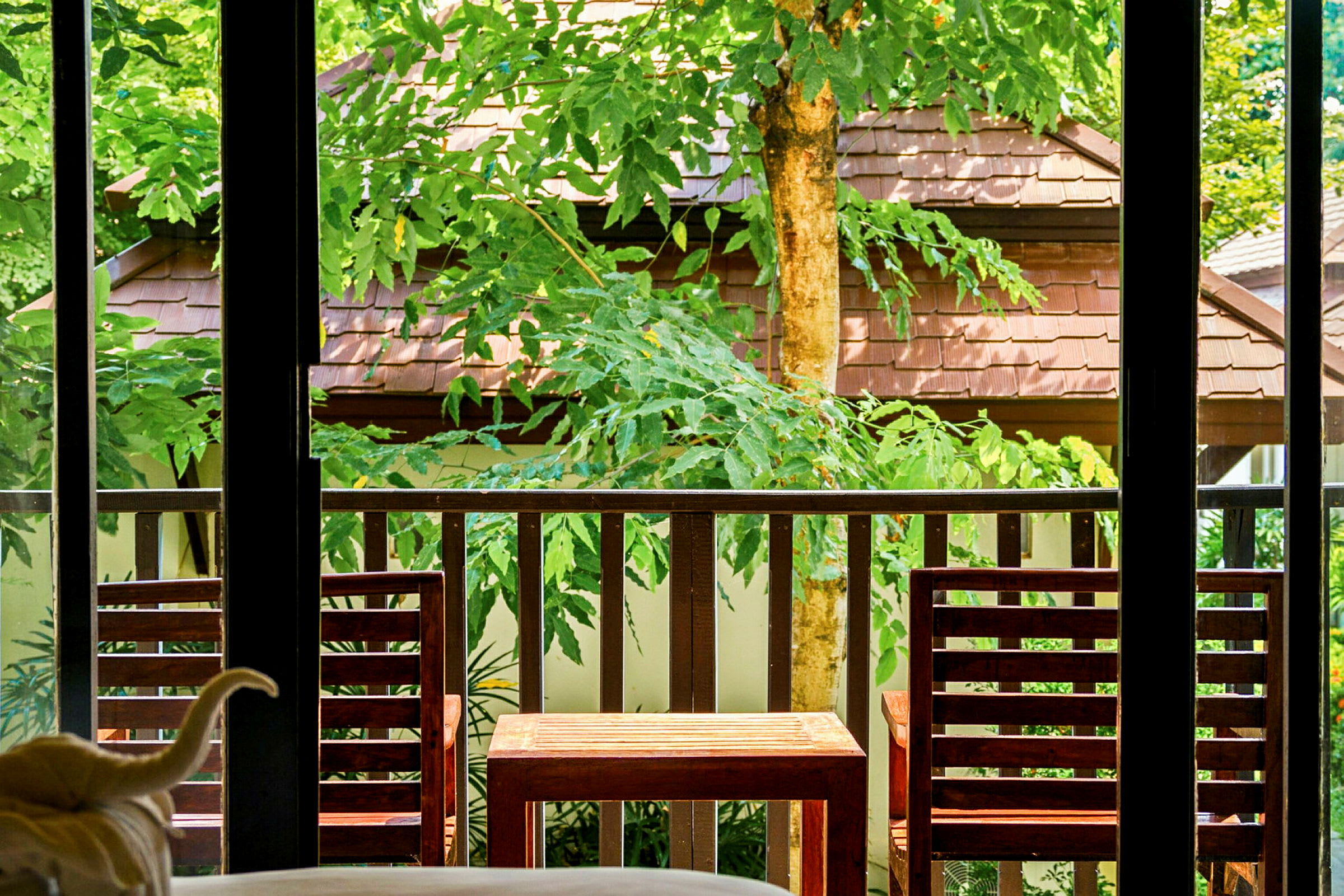 Utsikt fra et rom mot en terrasse med to tremøbler, omgitt av grøntområder og med taket på en bygning synlig i bakgrunnen. (AI-generert tekst)