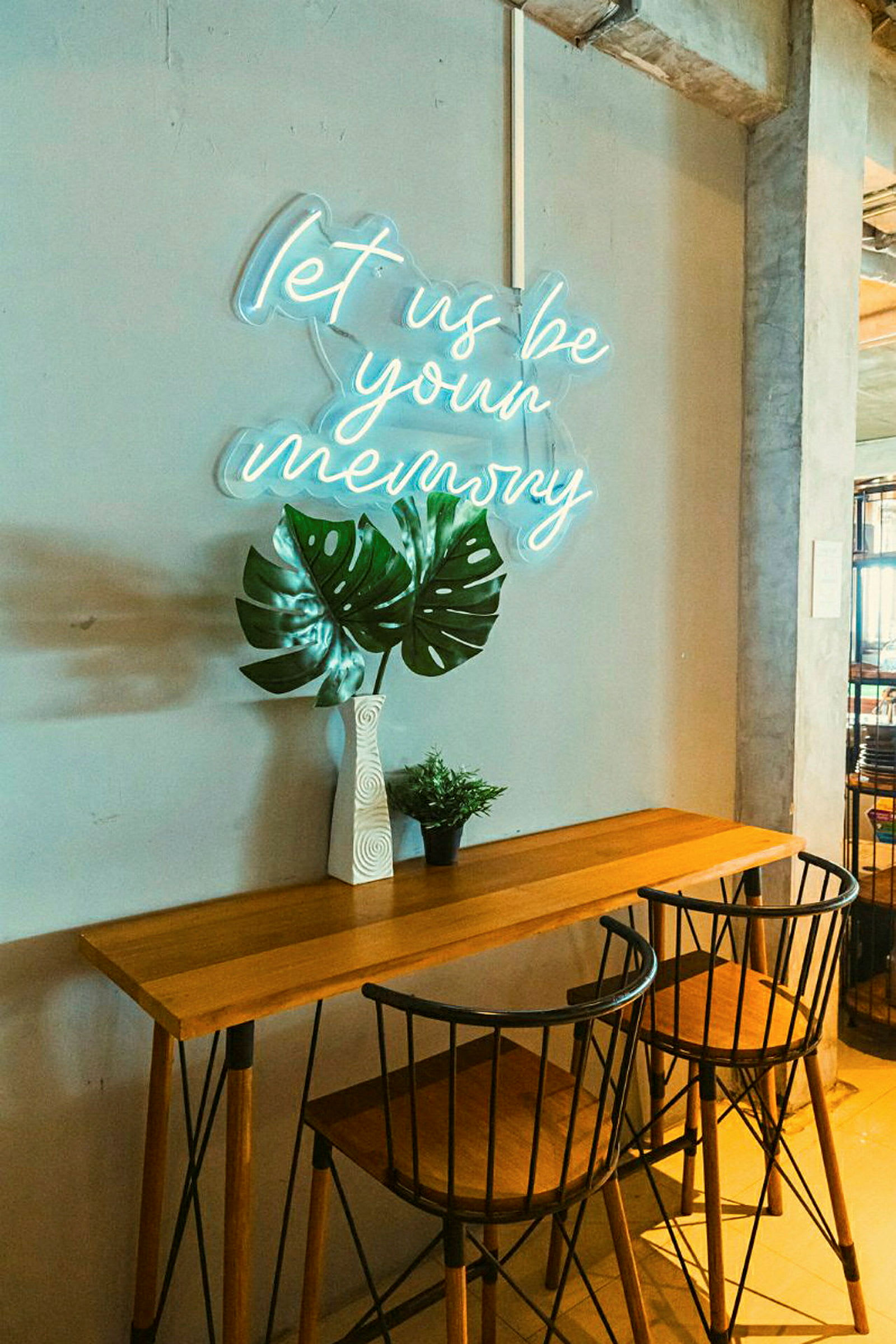 Et bord med to stoler, en vase med store grønne blader og en liten grønn plante, foran en vegg med et neonskilt som lyser opp teksten 'let us be your memory'. (AI-generert tekst)
