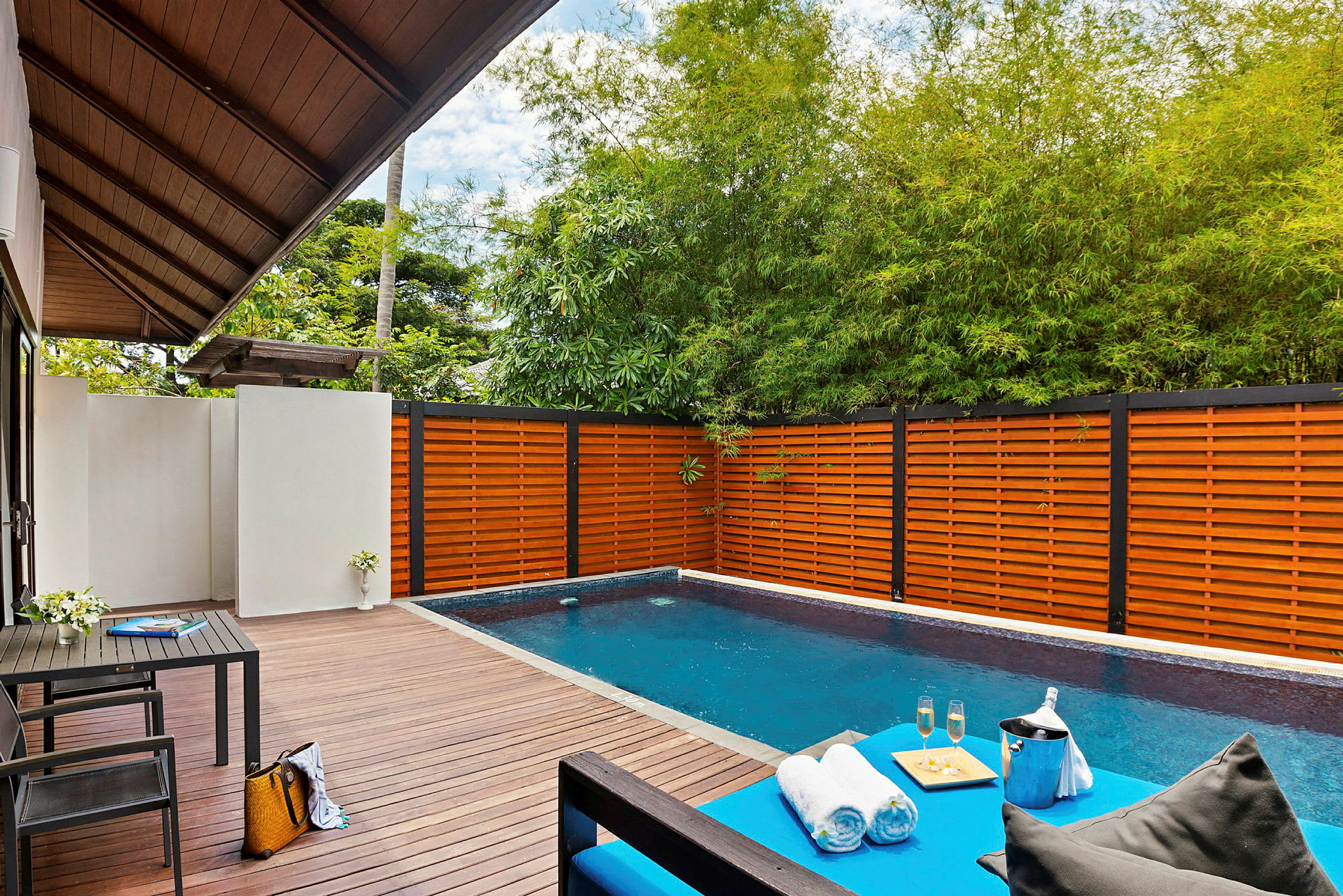 Et privat basseng med terrassebord, omgitt av gjerde i tre og grønne planter. En lounge med håndklær, drikke og en serveringsbrett vises i forgrunnen. (AI-generert tekst)