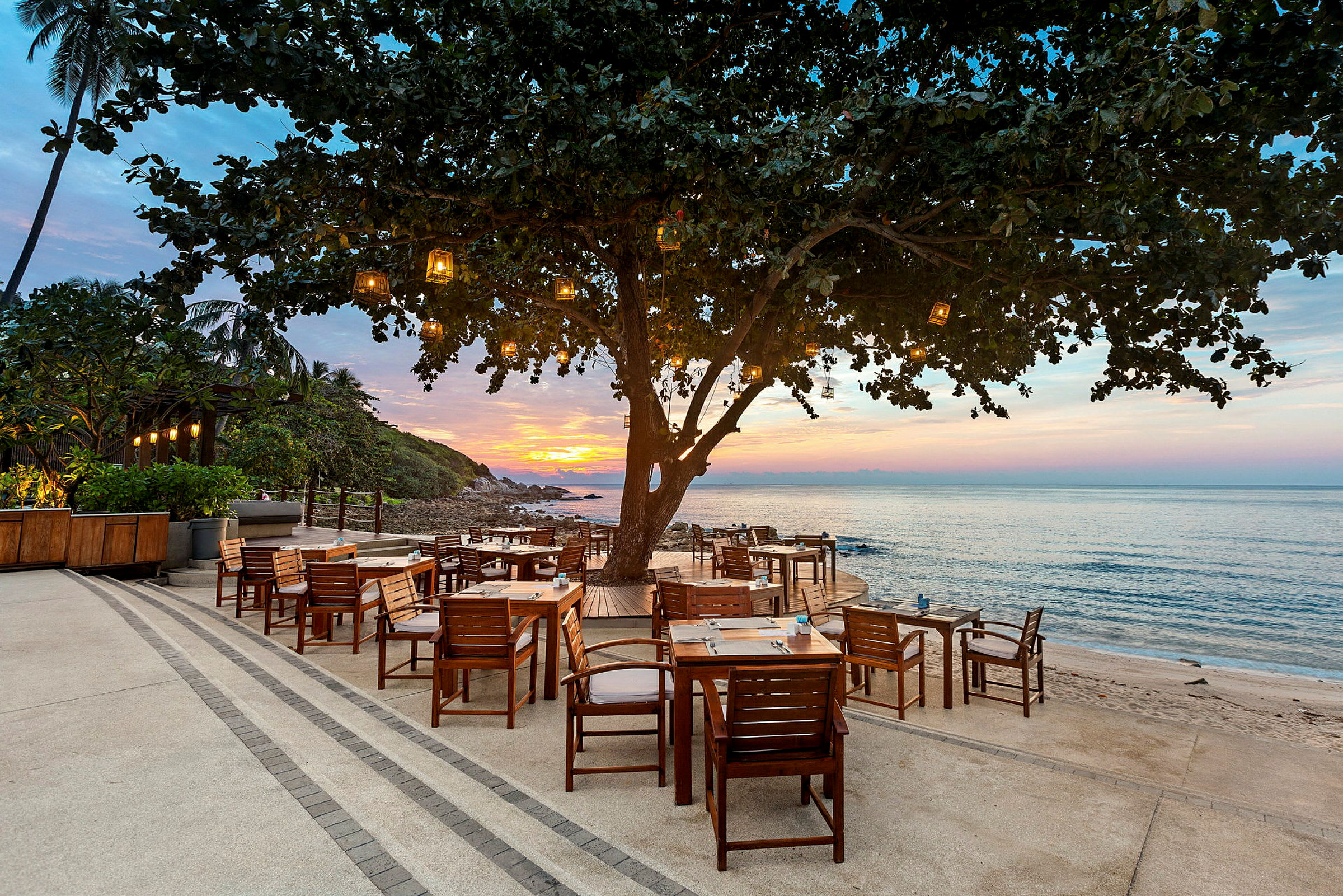 Utendørskafé med tremøbler på en terrasse ved stranden, opplyst av lykter med havet og solnedgangen i bakgrunnen. (AI-generert tekst)