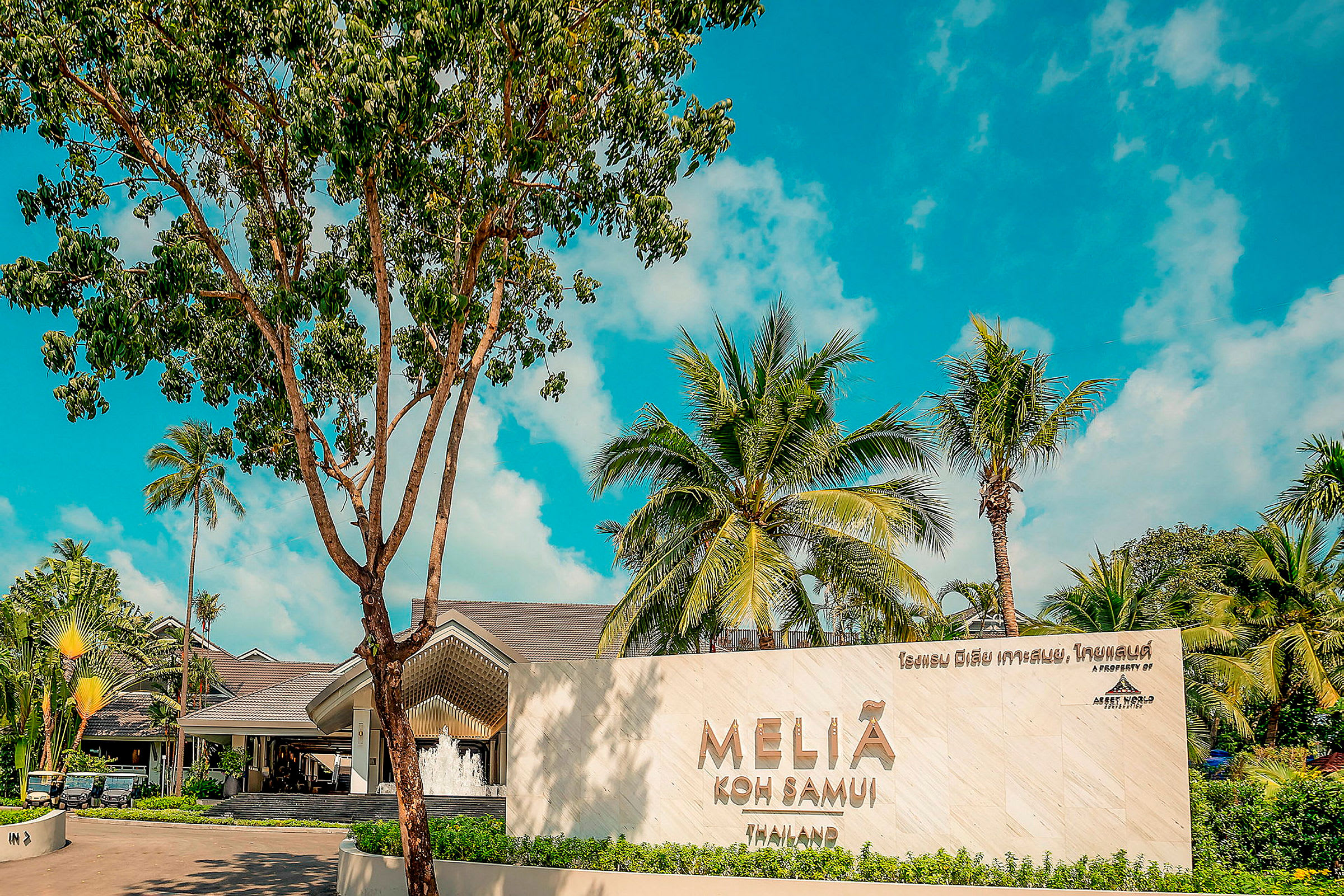 En tropisk resortinngang med et skilt der det står "Melia Koh Samui", omgitt av palmer og frodig vegetation med en blå himmel i bakgrunnen. (AI-generert tekst)