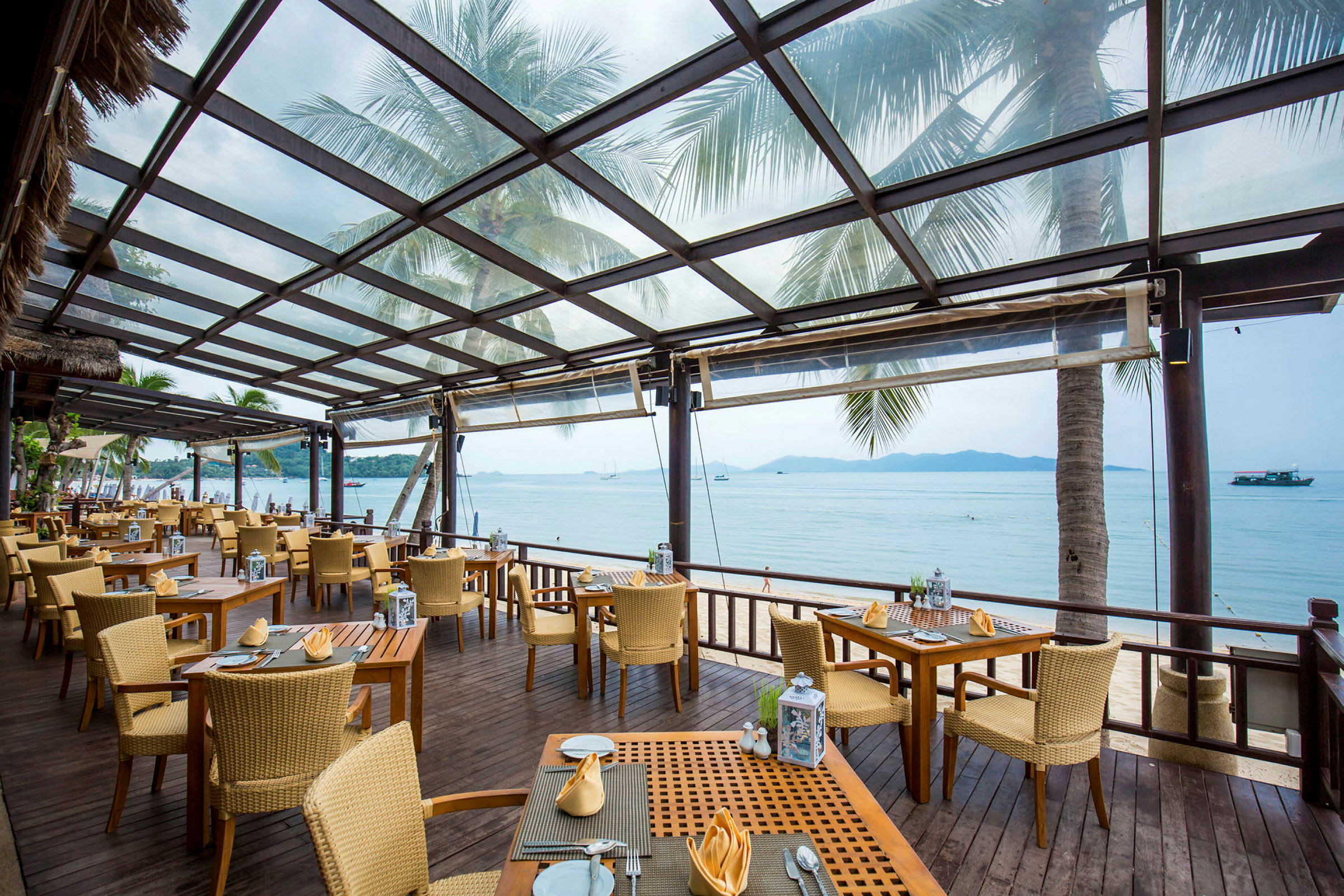 En restaurant med uteservering under et tak av glass. Stoler og bord er plassert på en treterrasse med utsikt over stranden og havet. (AI-generert tekst)