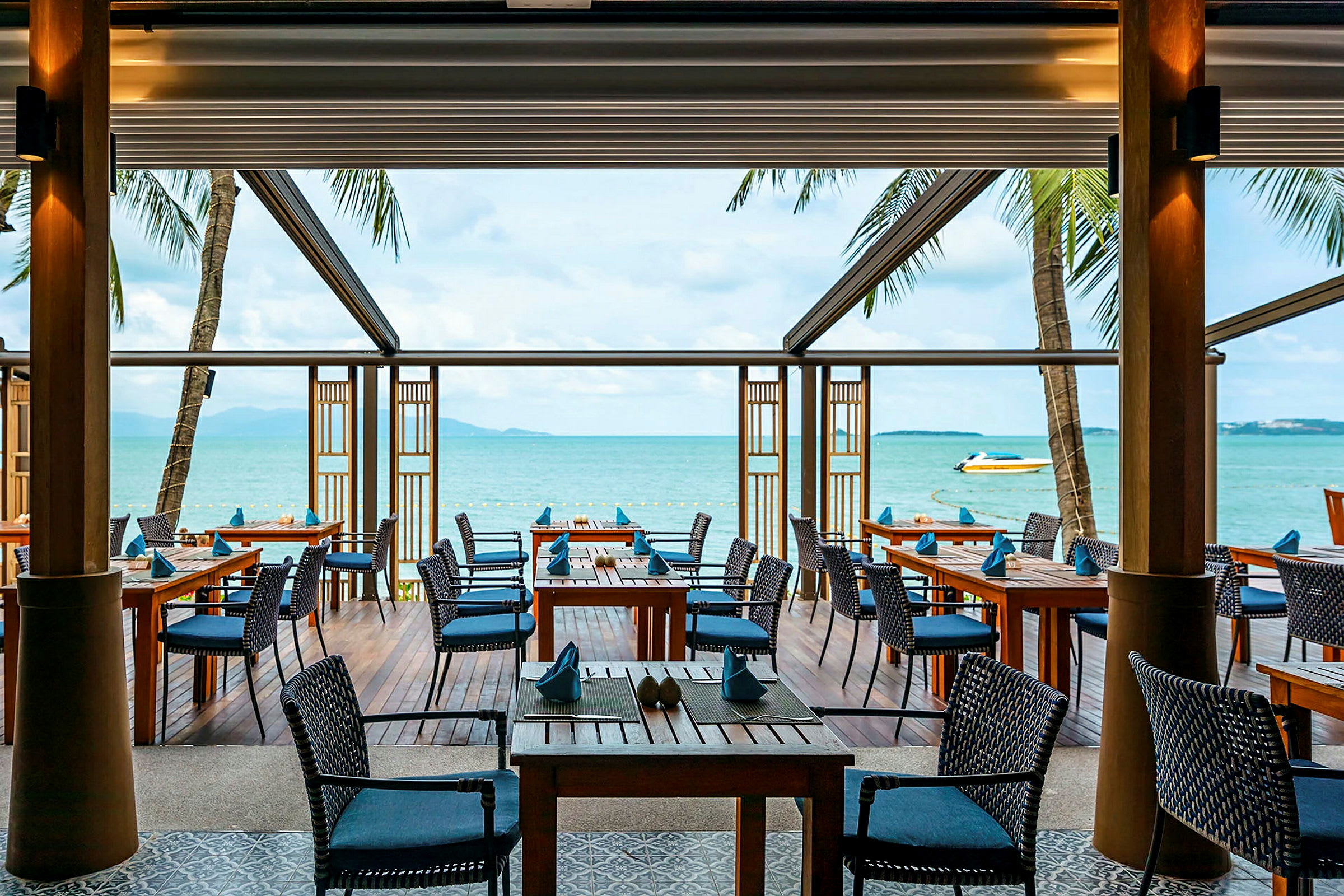 En uterestaurant med trebord og stoler ligger ved havet og tilbyr utsikt over vannet og en båt. (AI-generert tekst)