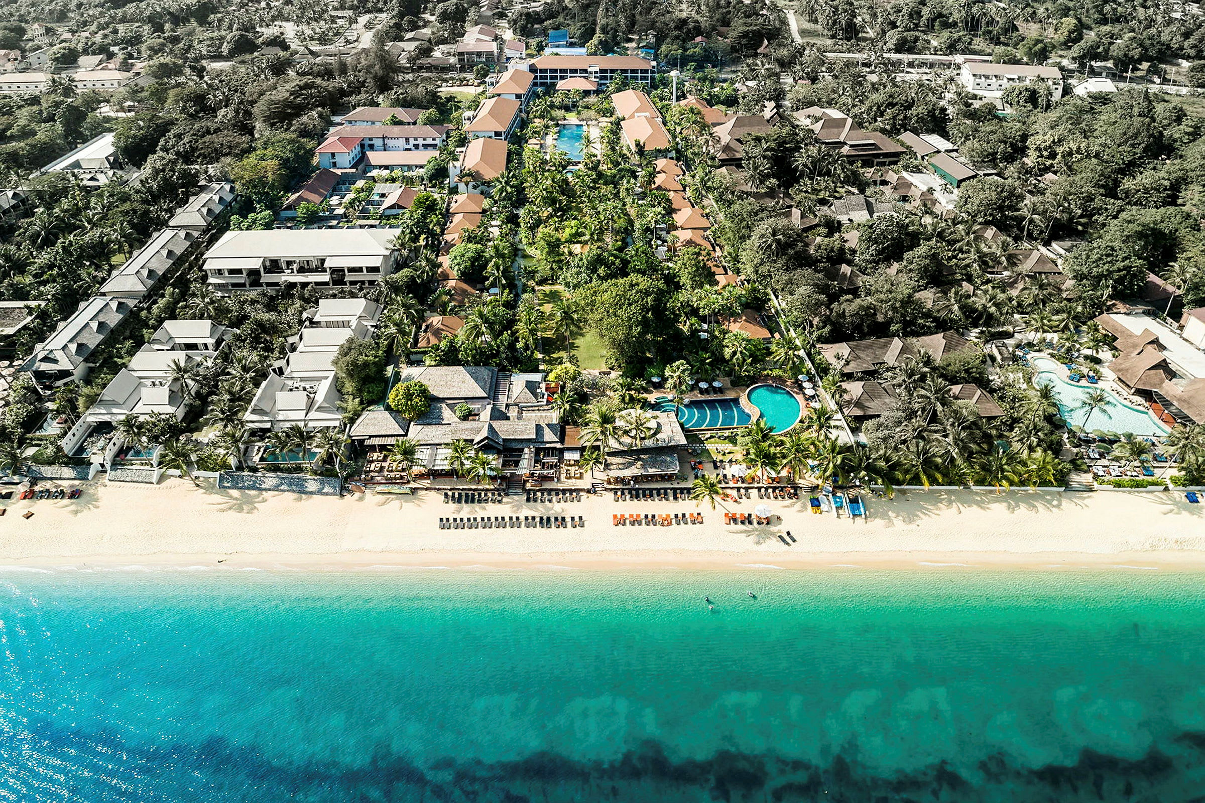 Luftfoto av en strand med turkist vann, solsenger og resortbygninger omgitt av grøntområder. (AI-generert tekst)