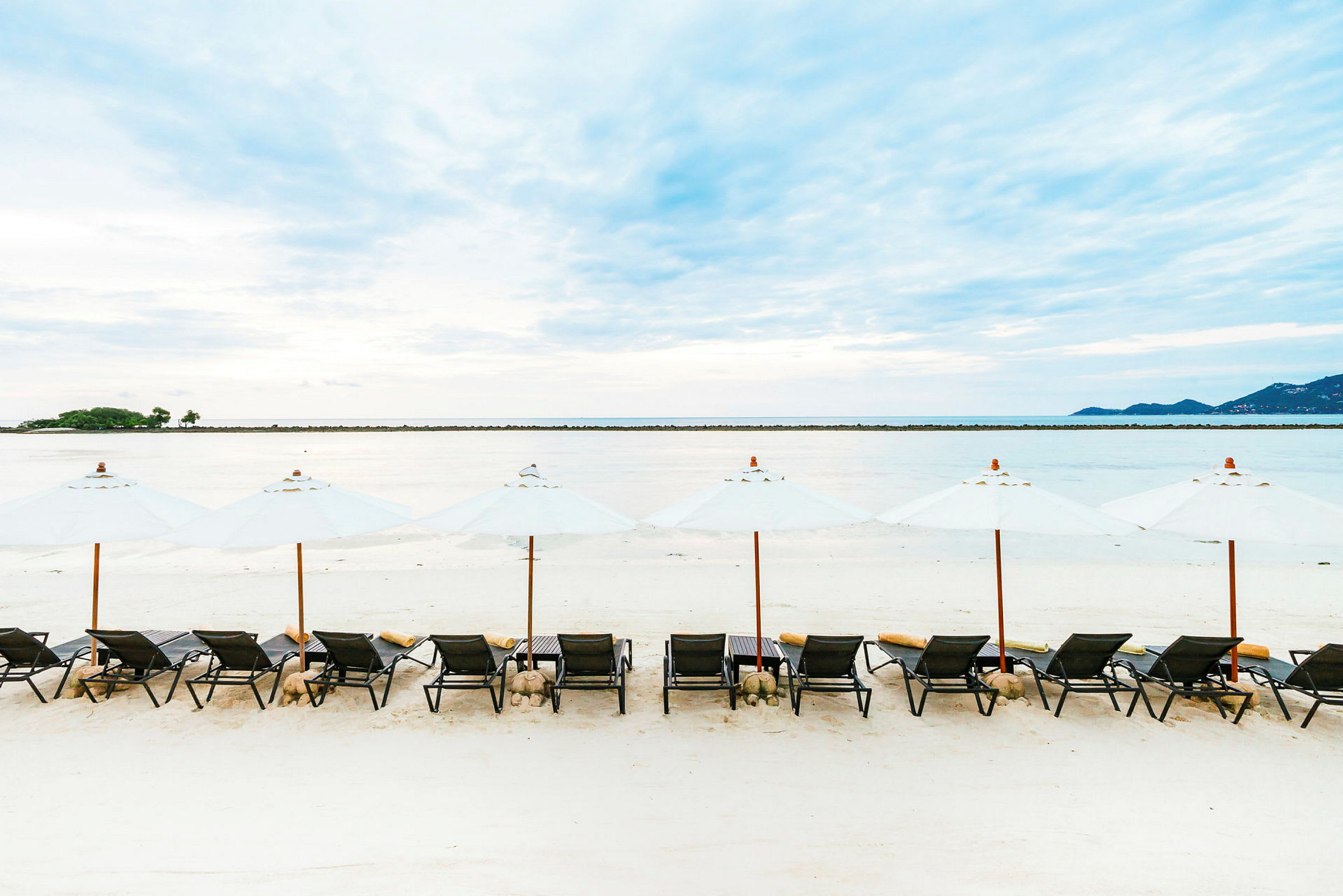 Bilde av en strand med solsenger og hvite parasoller plassert på sanden. En rolig vannflate og en horisontlinje er synlig i bakgrunnen. (AI-generert tekst)