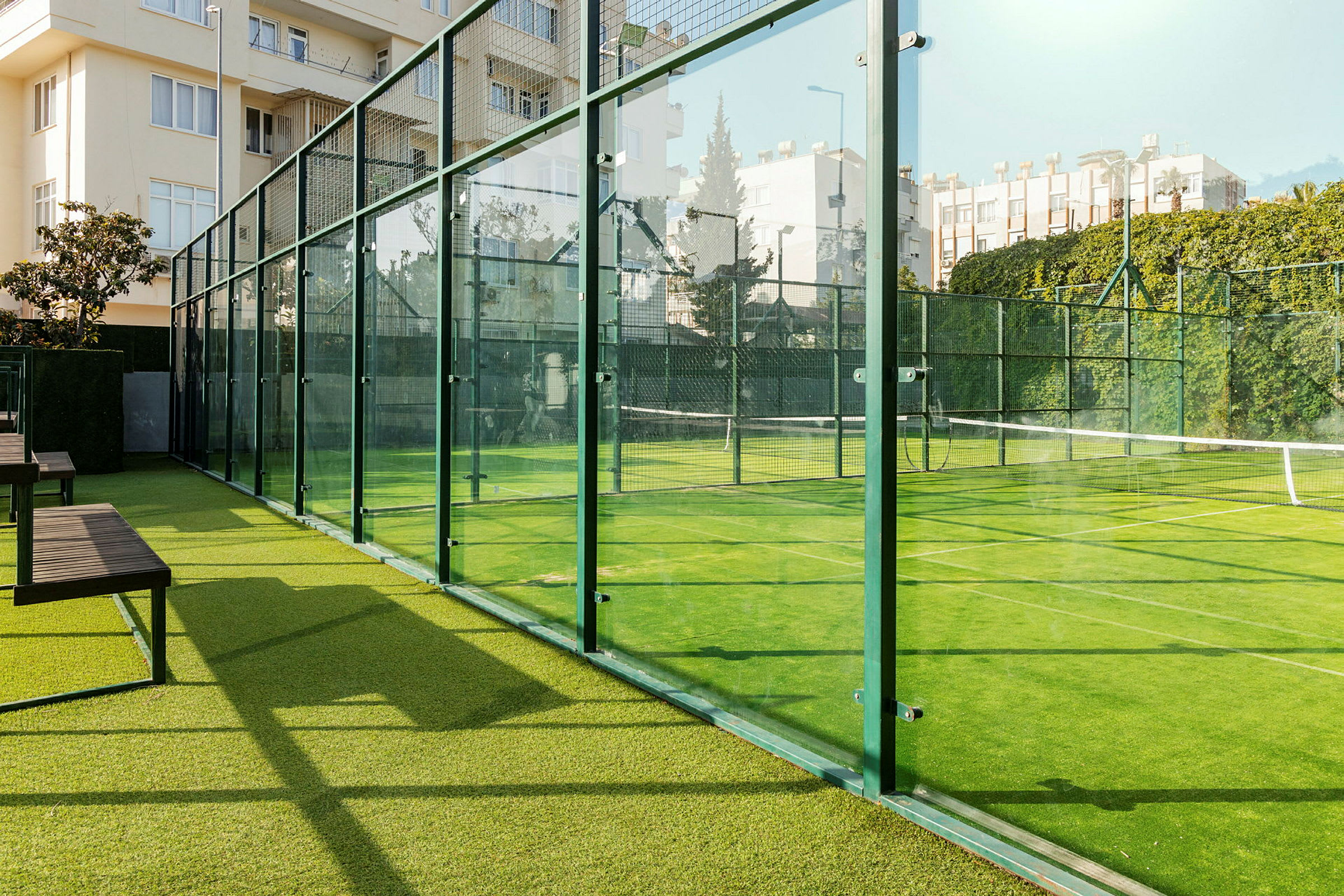 En utendørs padelbane omgitt av glassvegger, med grønn kunstgressdekke og omkringliggende bygninger. (AI-generert tekst)
