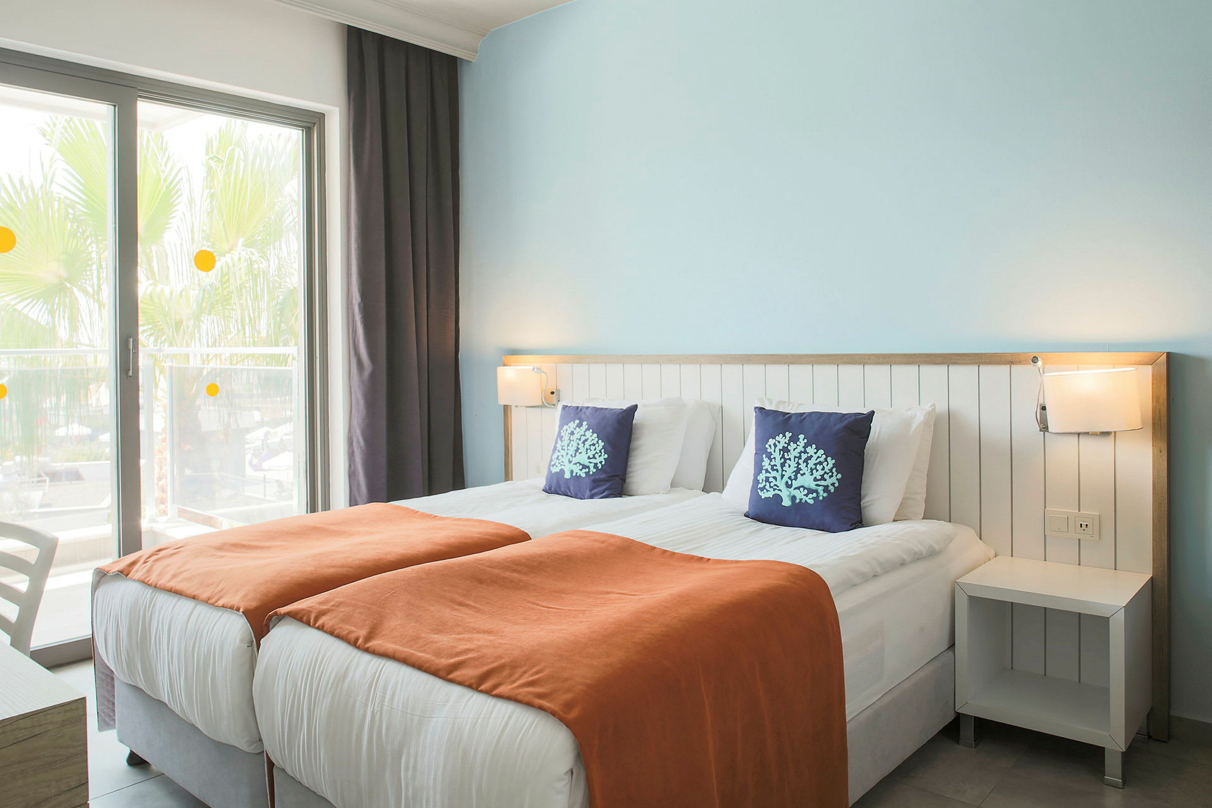 Hotellrom med to enkeltsenger, oransje tepper og dekorative puter. (AI-generert tekst)