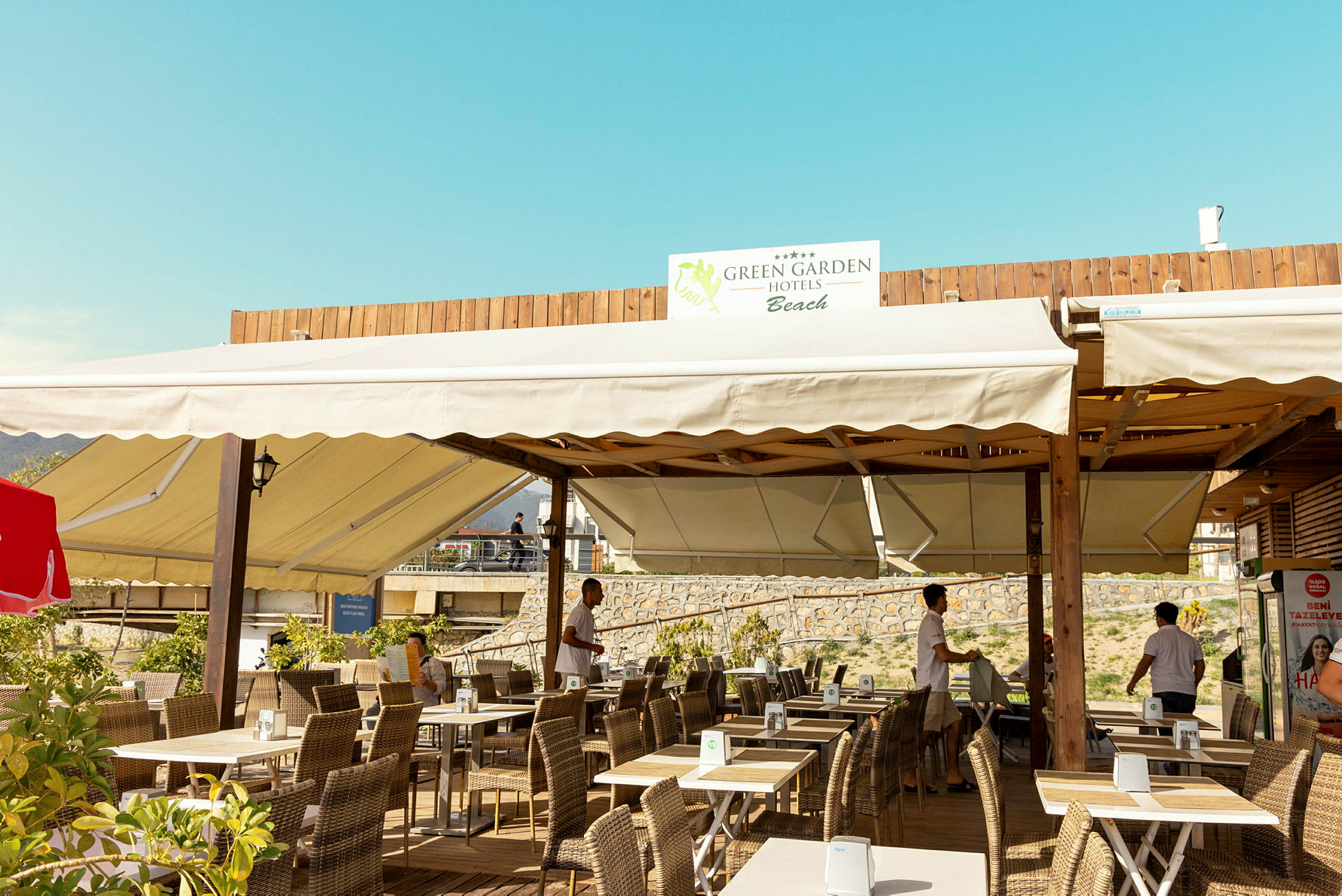 En uteservering med bord og stoler, med overliggende seiltak og et skilt med teksten 'Green Garden Hotels Beach'. (AI-generert tekst)