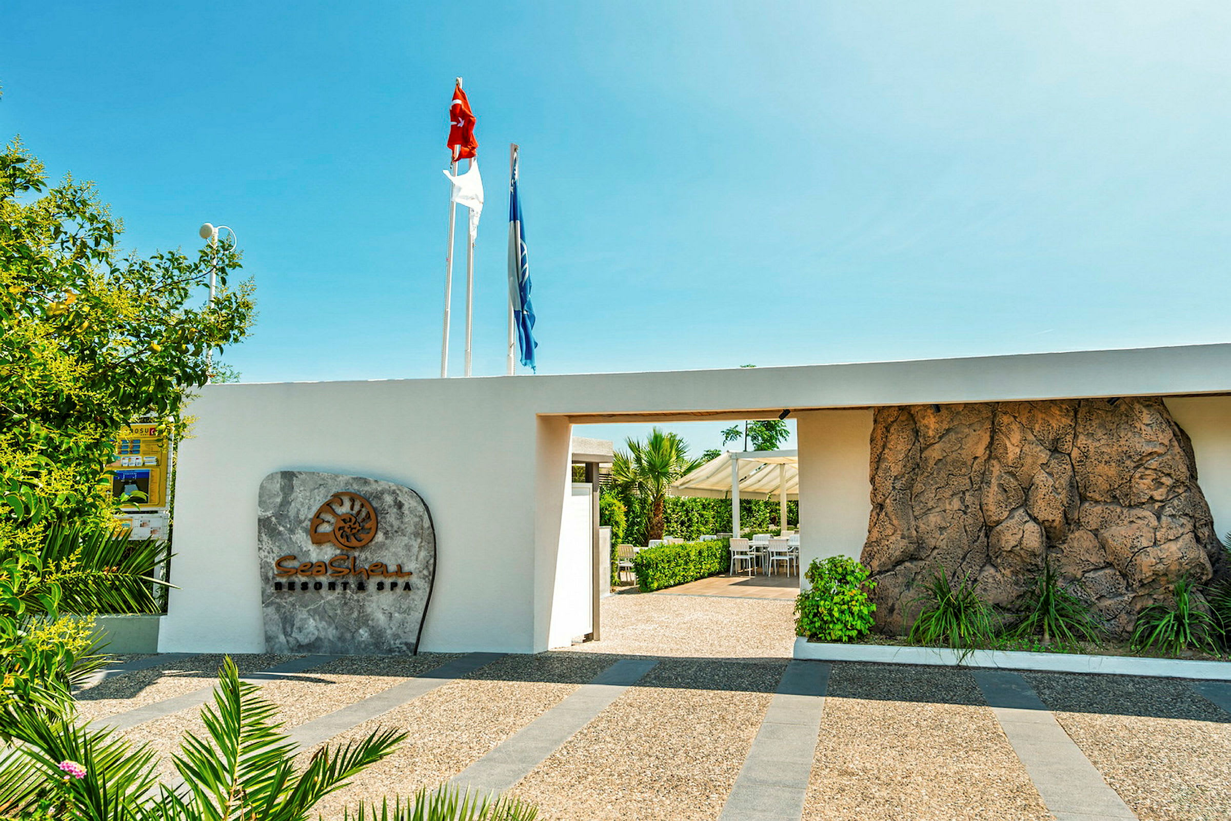 Inngang til en resort med et skilt som viser navnet 'Sea Shell Resort & Spa', omgitt av grønne omgivelser og tydelige flagg over. (AI-generert tekst)