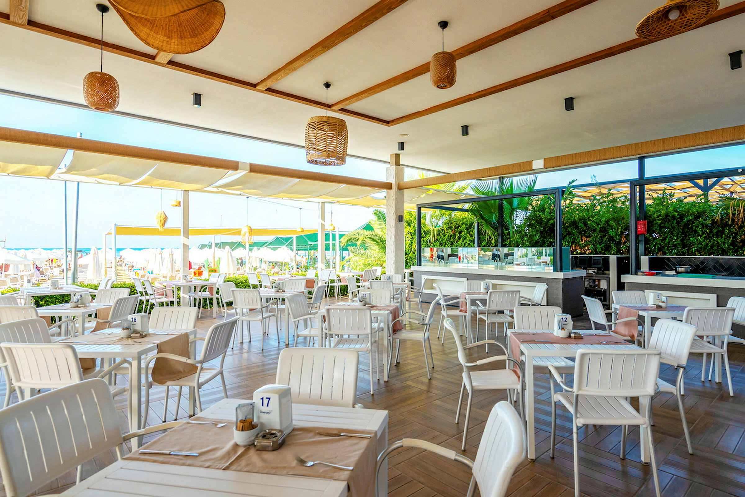 En utendørsrestaurant med bord og stoler i en lys innredning. I bakgrunnen er det grøntområder og utsikt over stranden. (AI-generert tekst)