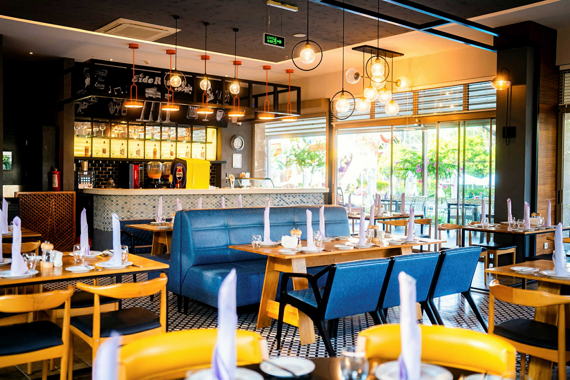 Et moderne innredet restaurantområde med bord, blå benkseter og hengende lamper i taket. (AI-generert tekst)