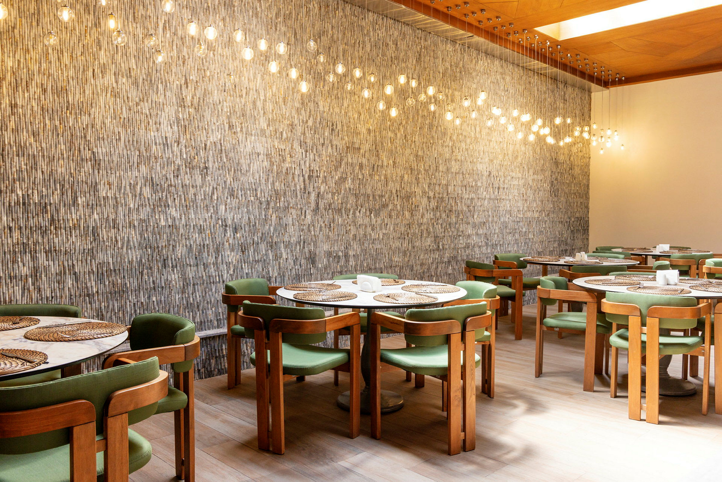 En innendørs restaurant med trestoler og grønne seter, runde bord med sivmatter og hengende lys med glødepærer over. (AI-generert tekst)