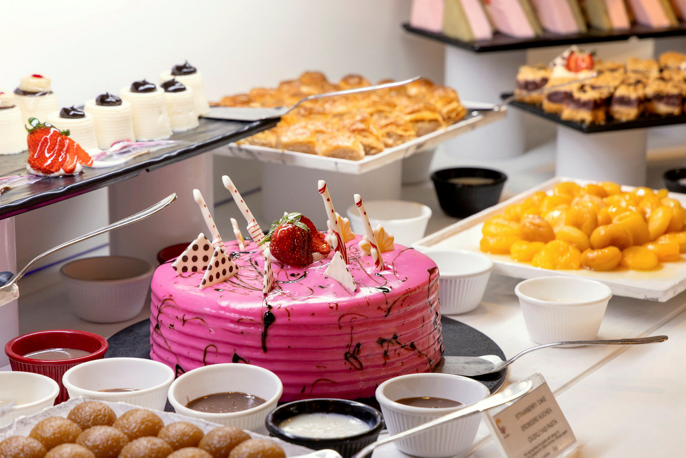 Et bord med forskjellige desserter inkludert en rosa kake, bakverk og skåler med sjokoladesaus. (AI-generert tekst)