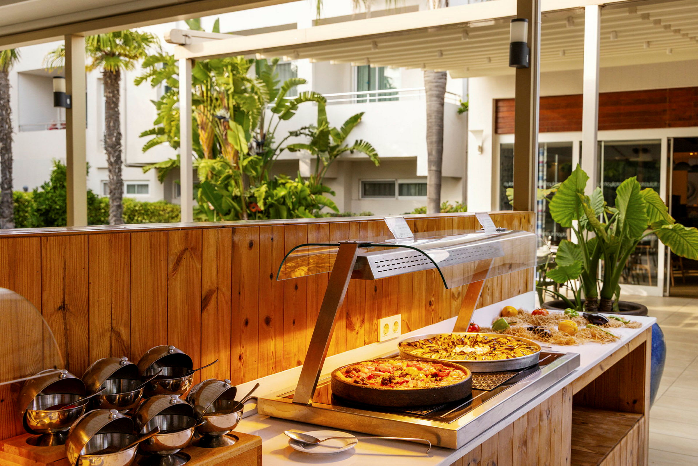 En buffet med mat, inkludert to pizzaer, omgitt av grønne planter og et utendørsmiljø. (AI-generert tekst)