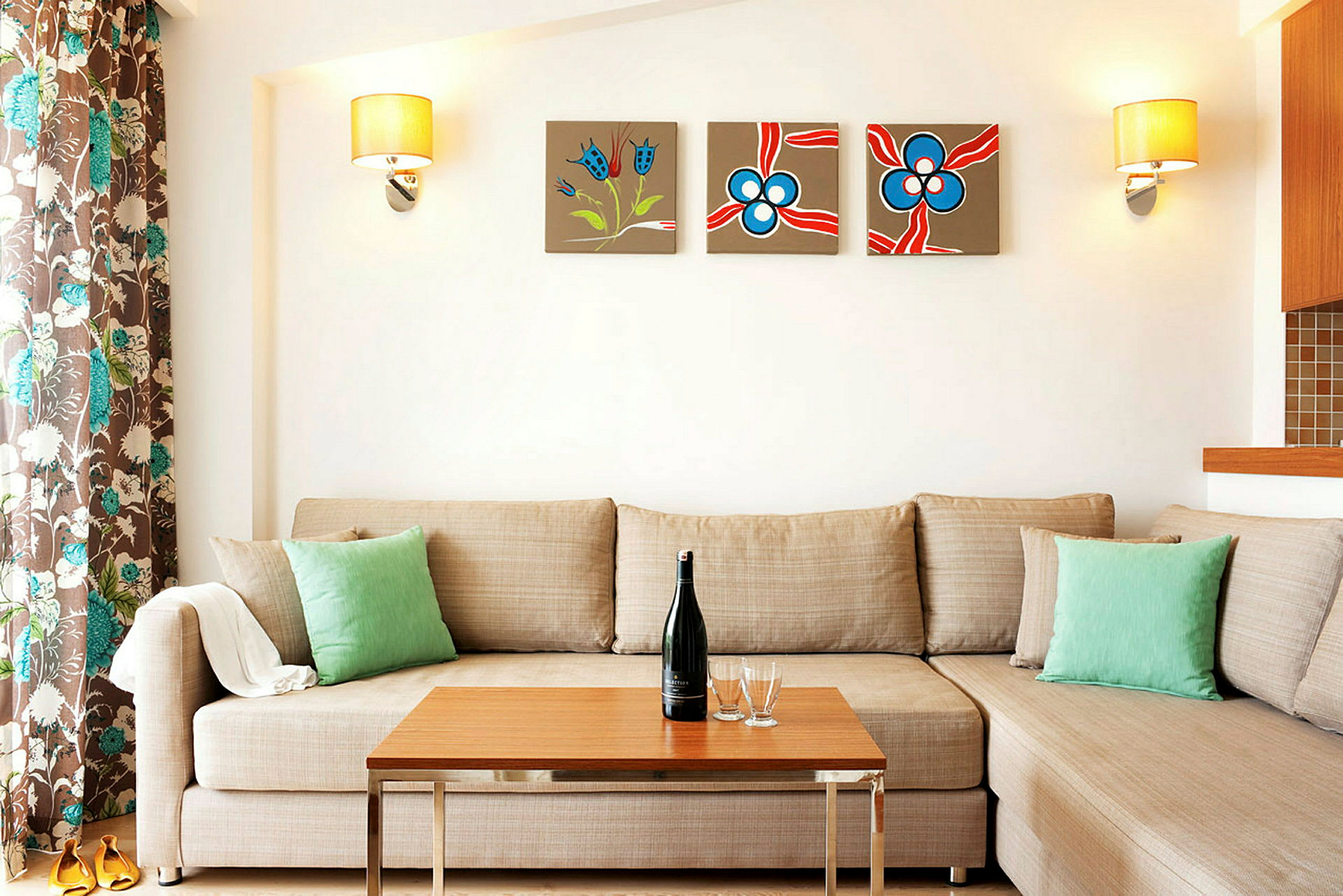 En beige sofa med grønne puter foran et trebord. Veggen bak har tre fargerike kunstverk og to lamper. (AI-generert tekst)