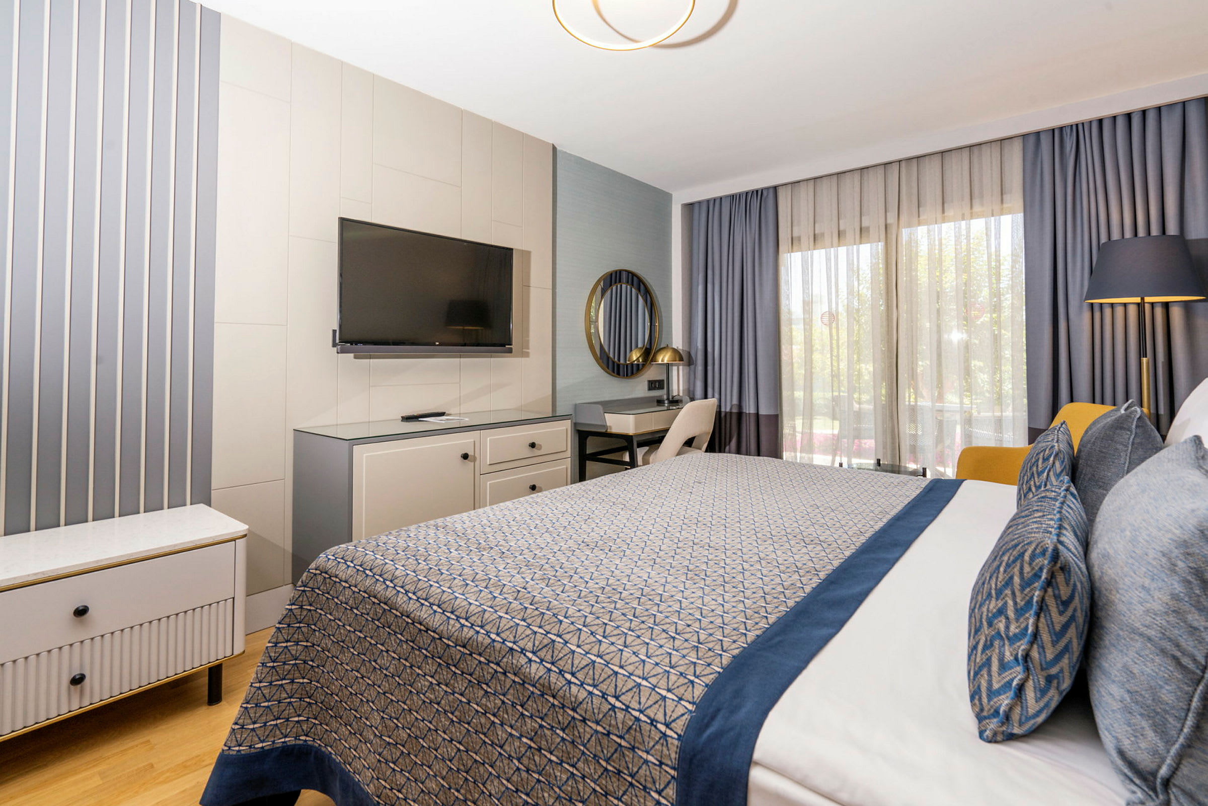 Hotellrom med moderne innredning, inkludert seng med blå detaljer, skrivebord, TV og store vinduer med gardiner. (AI-generert tekst)