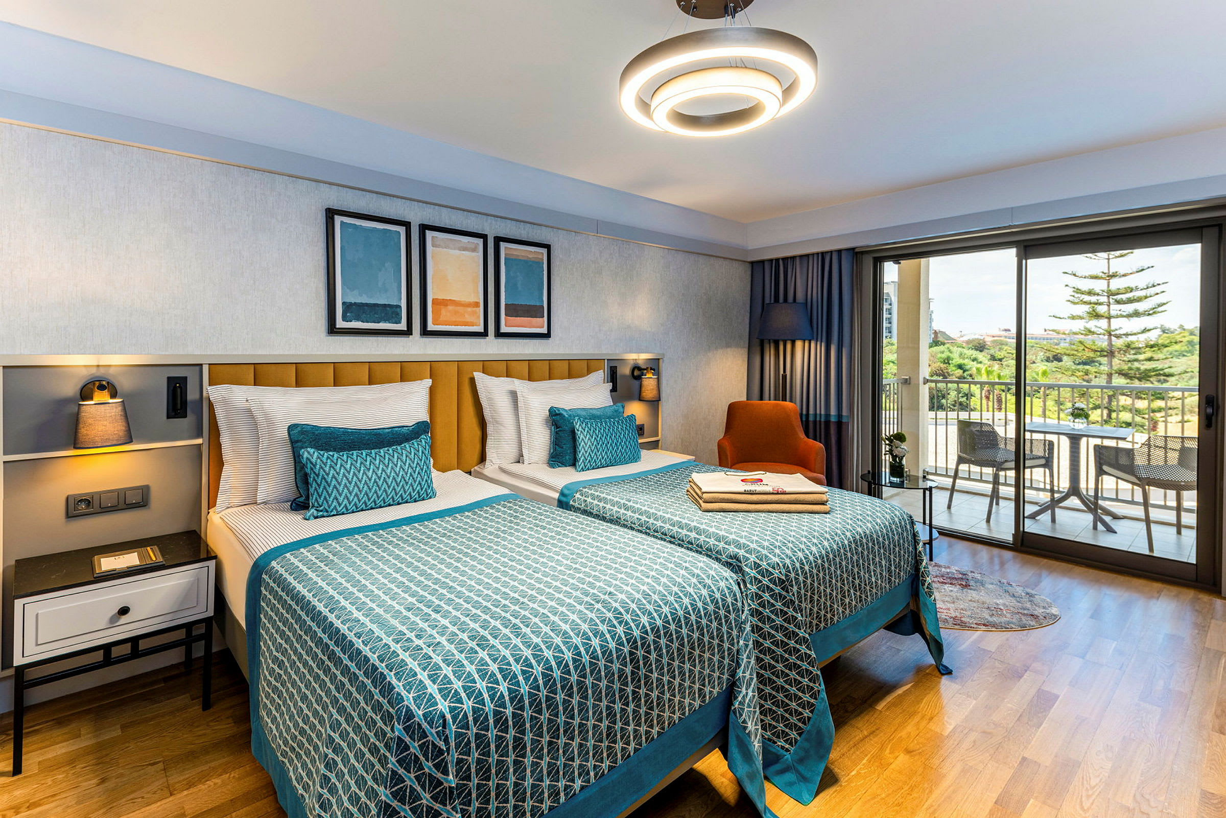Hotellrom med to senger ved siden av hverandre og dekorerte sengetepper, vindu med utsikt til en balkong utendørs. (AI-generert tekst)