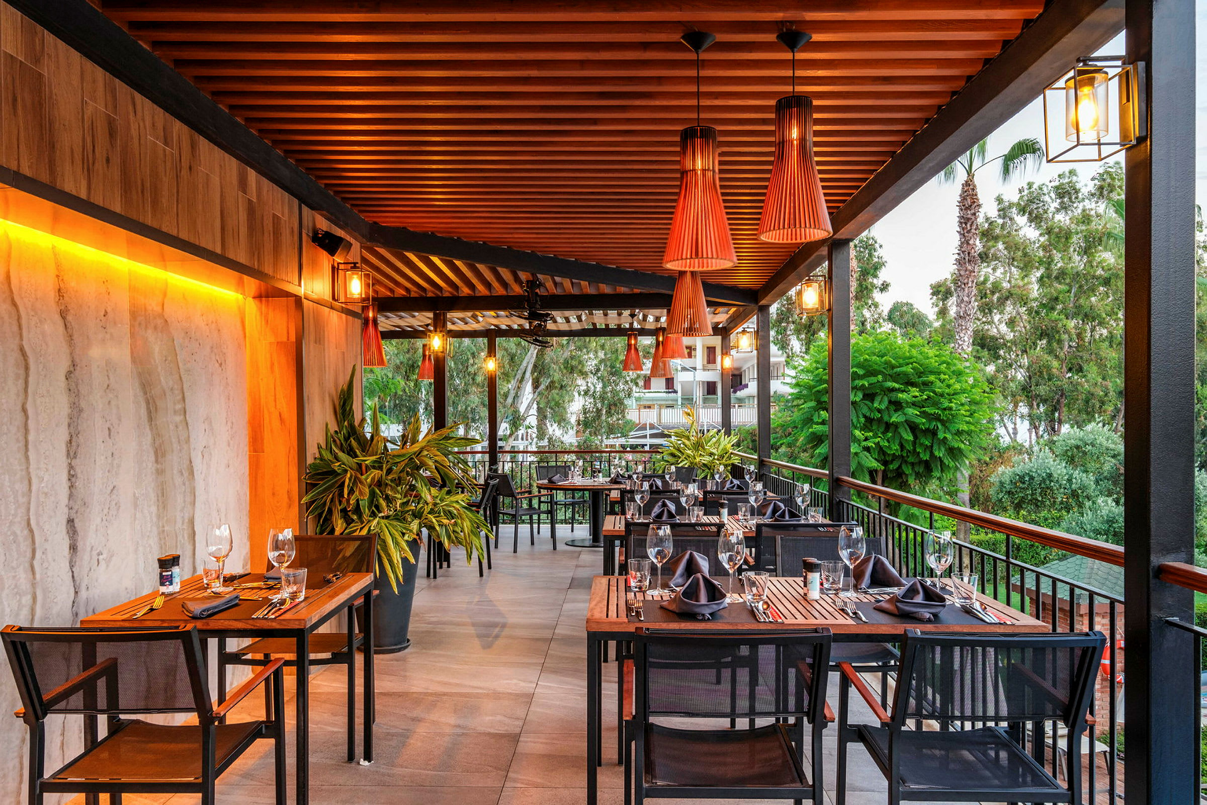 En terrasse med bord og stoler arrangert for middag, med grøntområder og dekorative lamper. (AI-generert tekst)