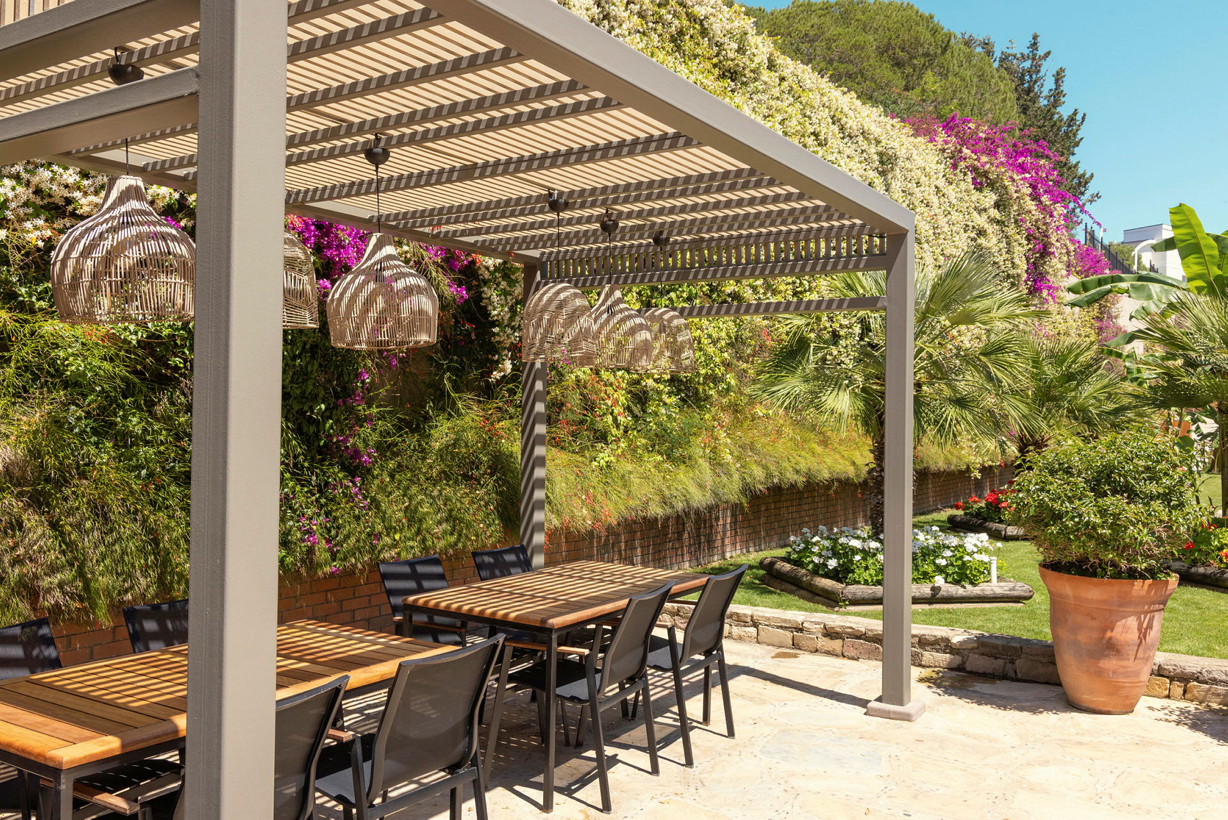 En utendørs sittegruppe med bord og stoler under en pergola, omgitt av grønt og blomster. (AI-generert tekst)