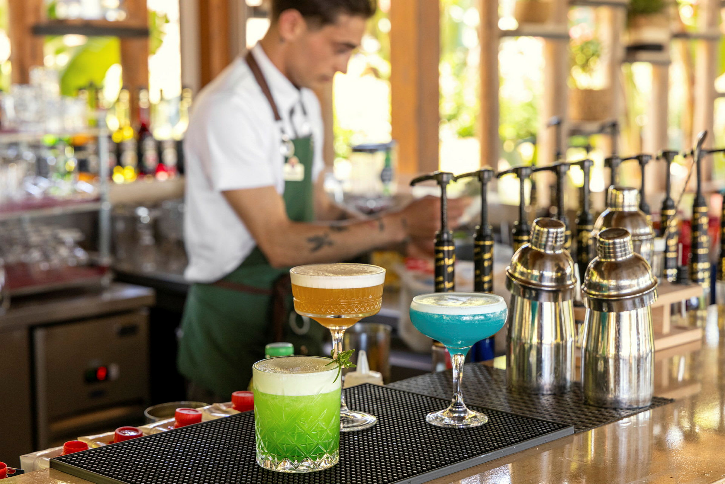 En bar med tre fargerike cocktails, shakers og en bartender i bakgrunnen. (AI-generert tekst)