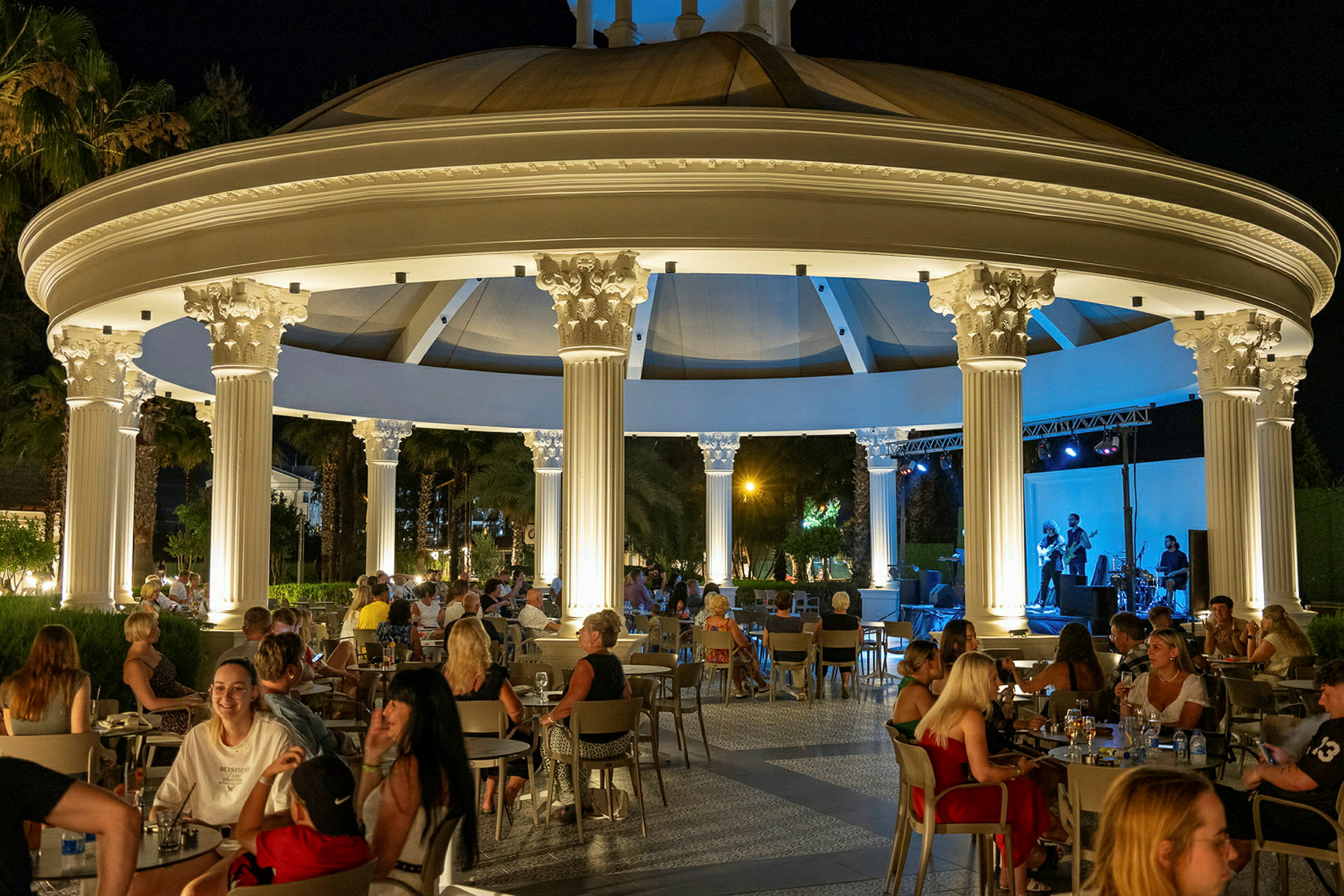 Utendørs restaurant med konsert på kveldstid, med belysning og folk ved bord. (AI-generert tekst)