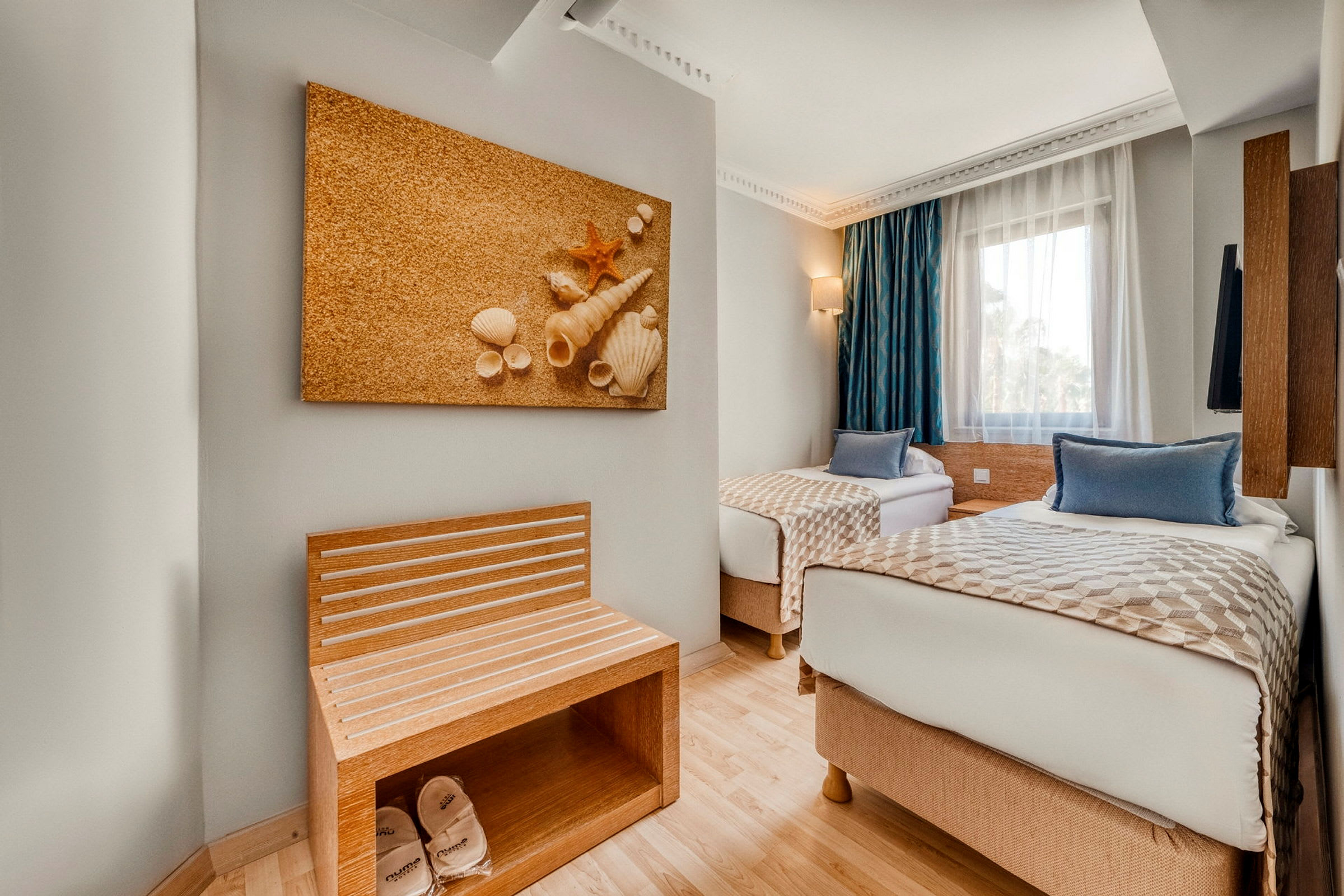 To enkeltsenger i et hotellrom med en veggdekorasjon av skjell og en benk med tøfler. Ved siden av er et vindu med blå gardiner. (AI-generert tekst)