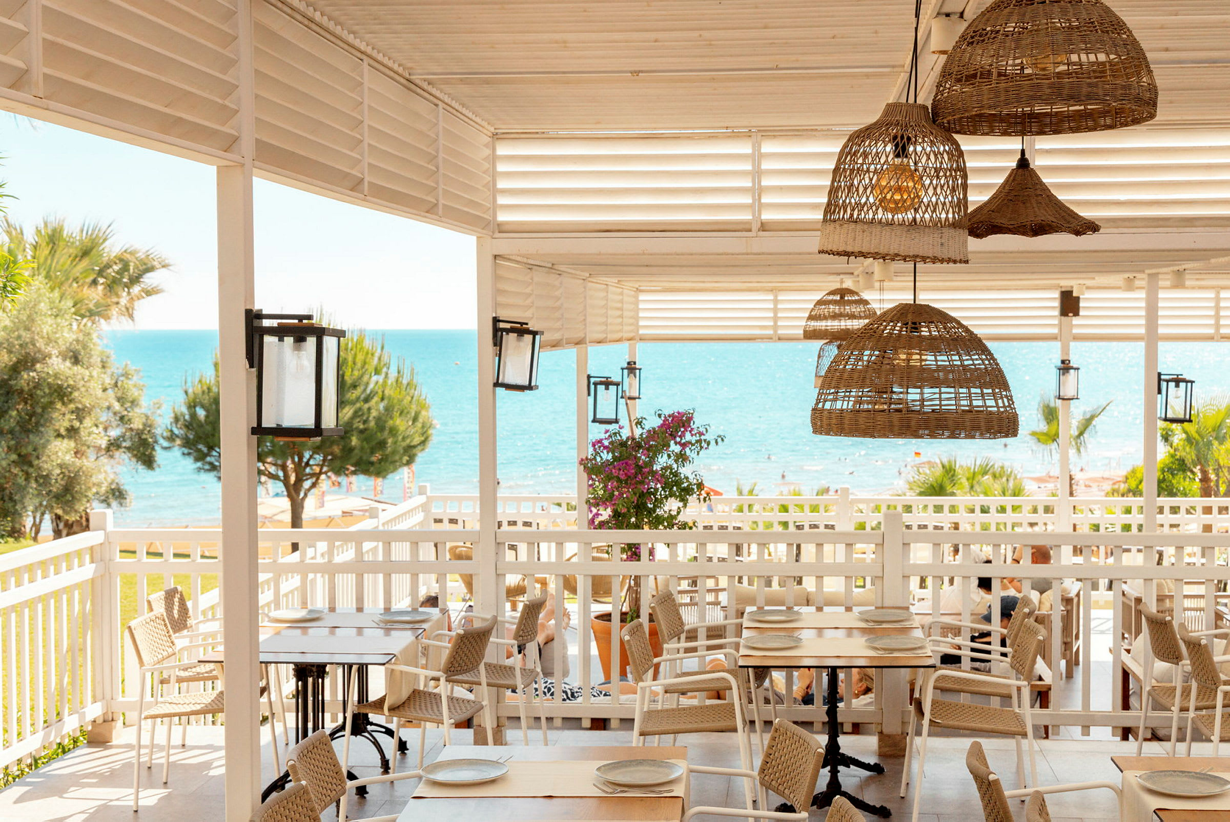 En strandrestaurant med trebord, rottingstoler og utsikt over havet. (AI-generert tekst)