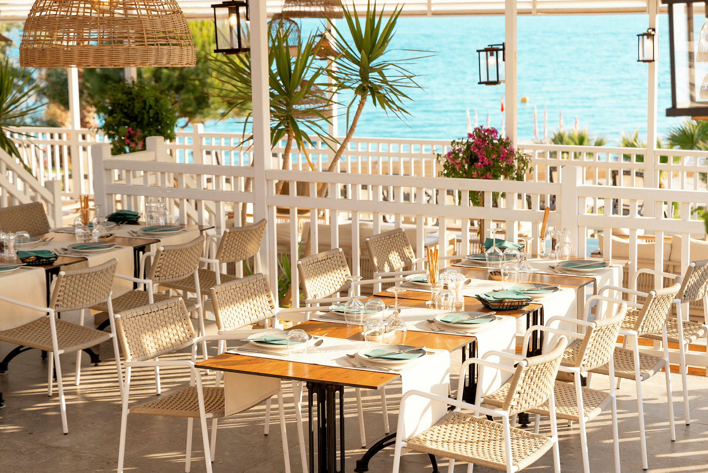 En utendørs restaurant med bord dekket med tallerkener og glass. Utsikt over havet i bakgrunnen. (AI-generert tekst)
