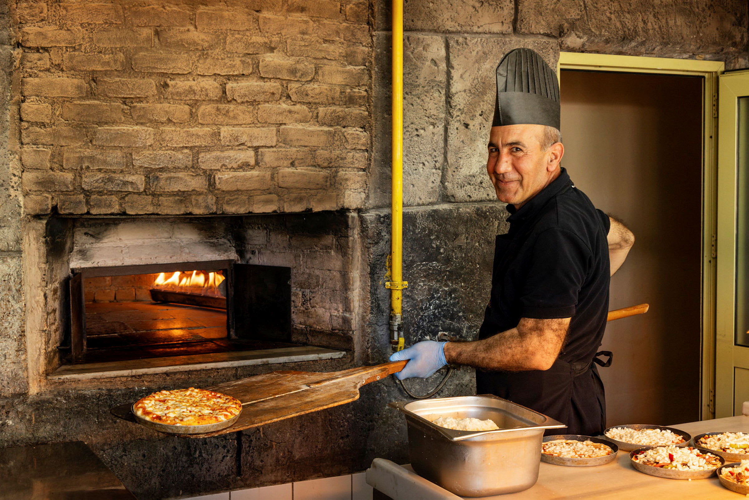En pizzabaker som tar ut en pizza fra en steinovn, klar til å serveres. (AI-generert tekst)