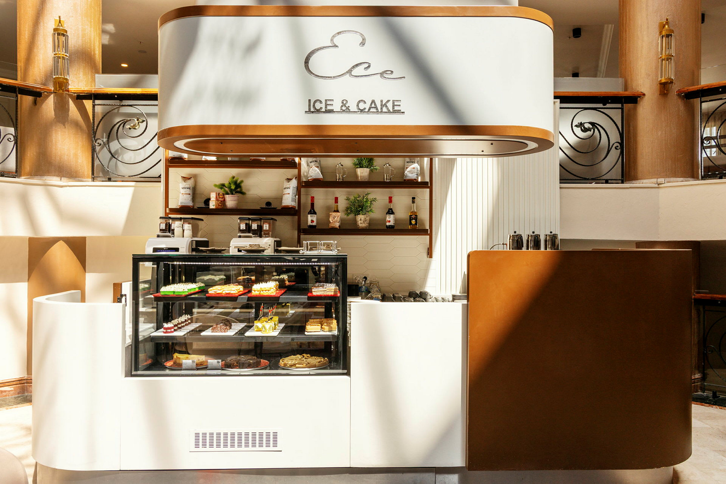 En kafé med glassdisk som viser ulike bakverk, skilt med teksten 'Ice & Cake' og innredning i lyse toner. (AI-generert tekst)