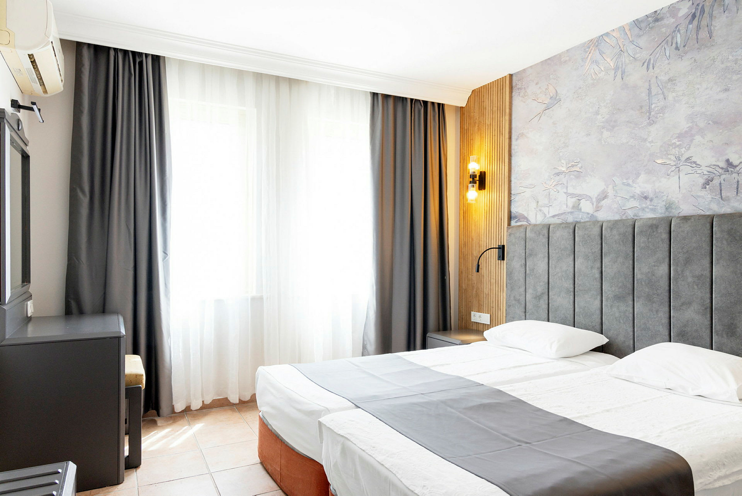 Et hotellrom med en dobbeltseng, grå overkast, vindu med lyse gardiner og veggpanel med dekorativt mønster. (AI-generert tekst)