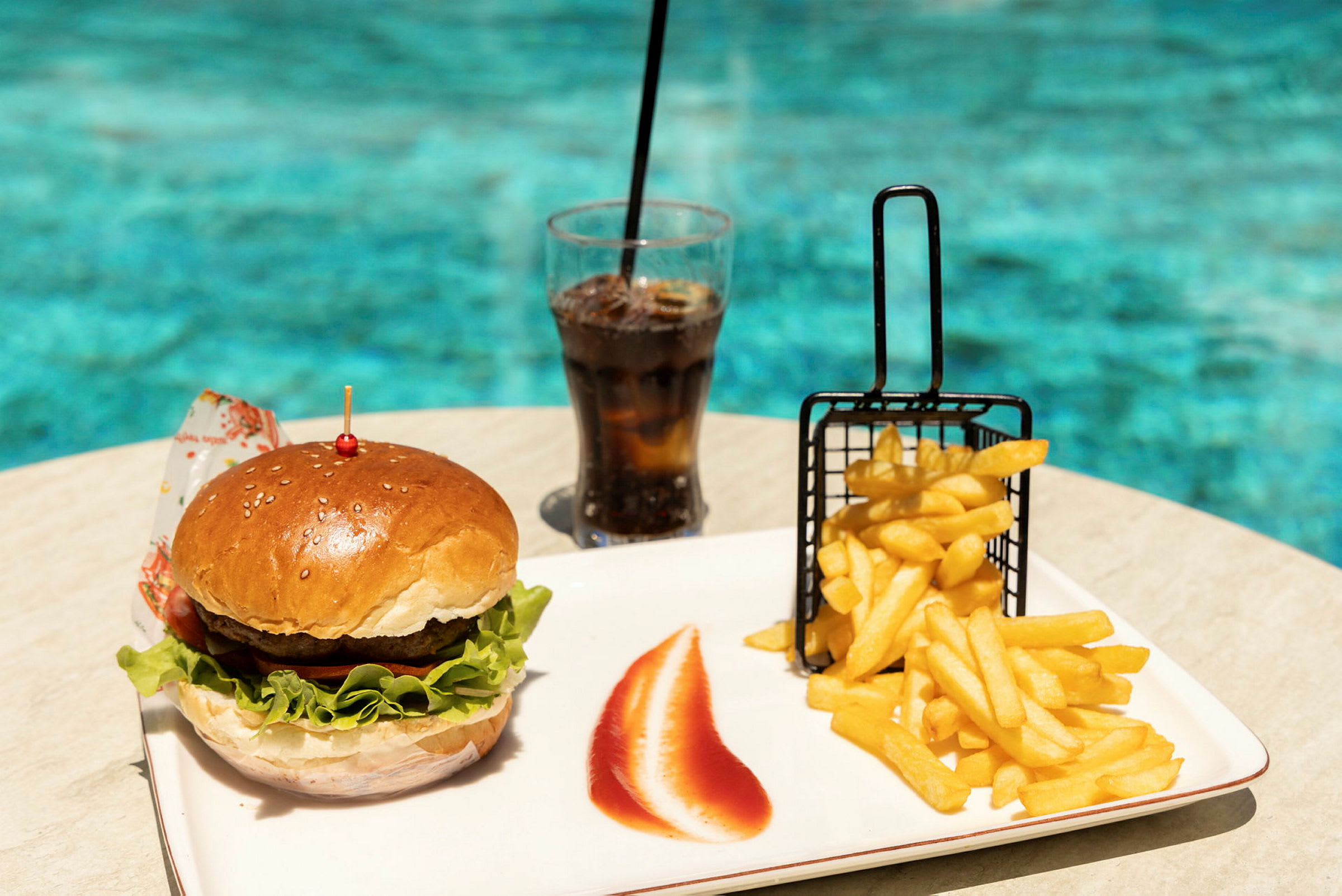 En hamburger, pommes frites og et glass brus på et bord ved et basseng. (AI-generert tekst)